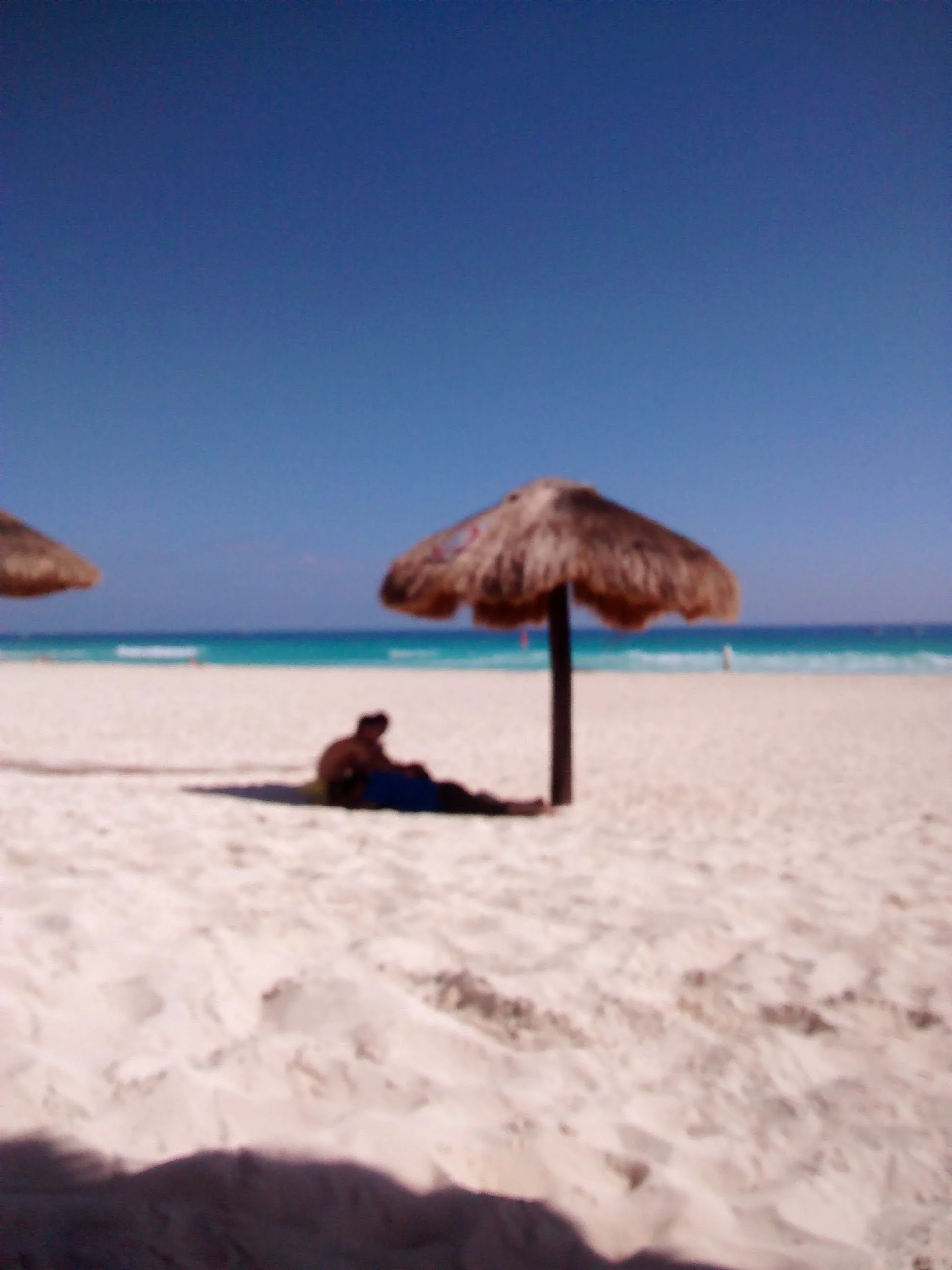 Beach scene with two straw umbrellas on sandy shore, person sitting under umbrella, calm ocean in background, clear blue sky.