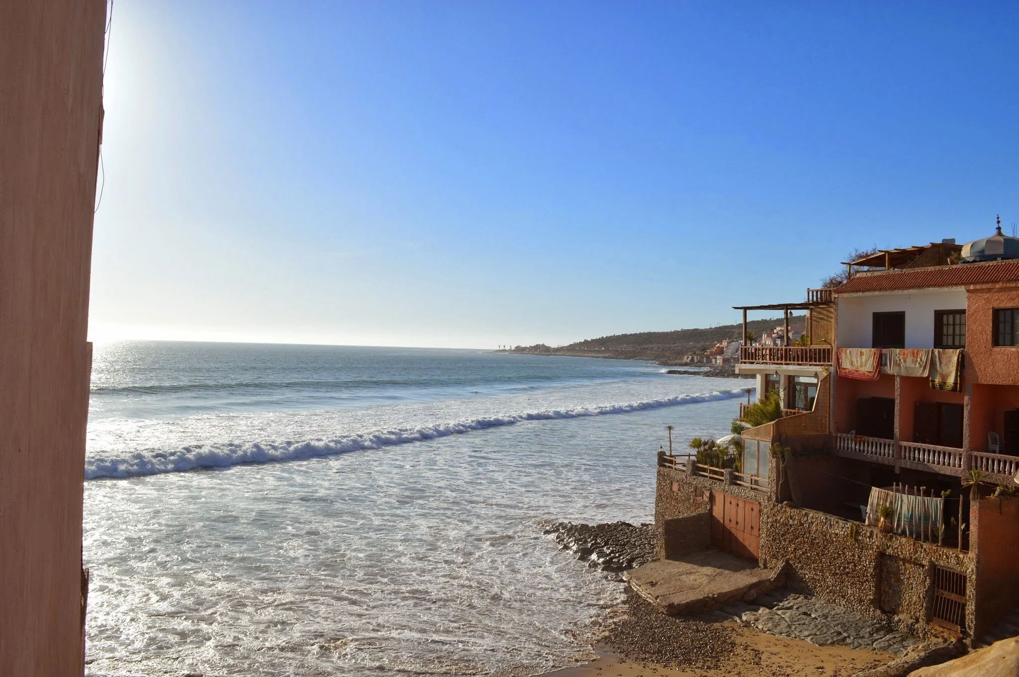 Beach view with ocean waves, sandy shore, and buildings with balconies overlooking the beach on a sunny day.
