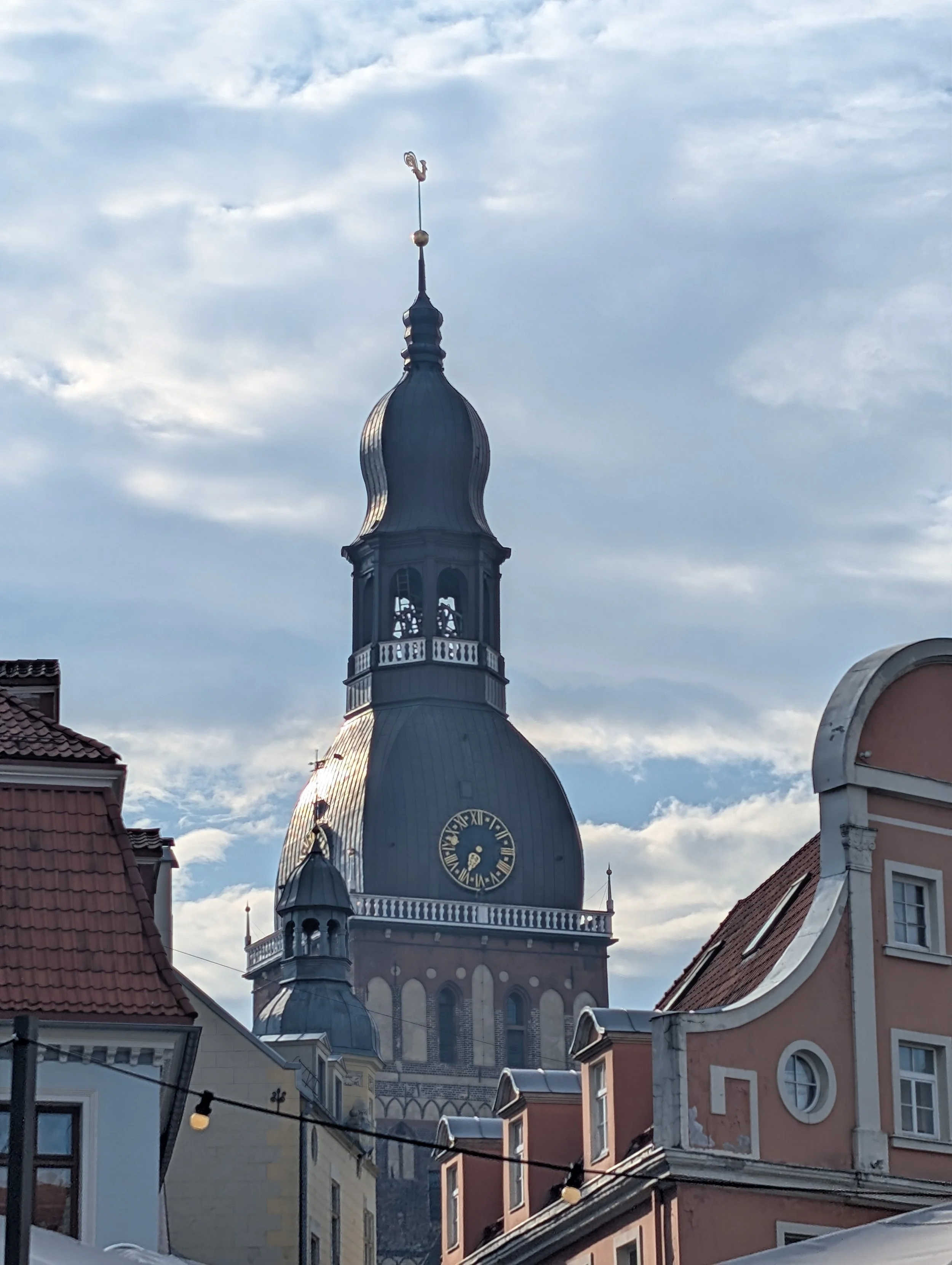 Church steeple with a clock, topped with a weather vane showing a rooster, against a cloudy sky. Surrounding buildings with tiled roofs and facades are visible in the foreground.