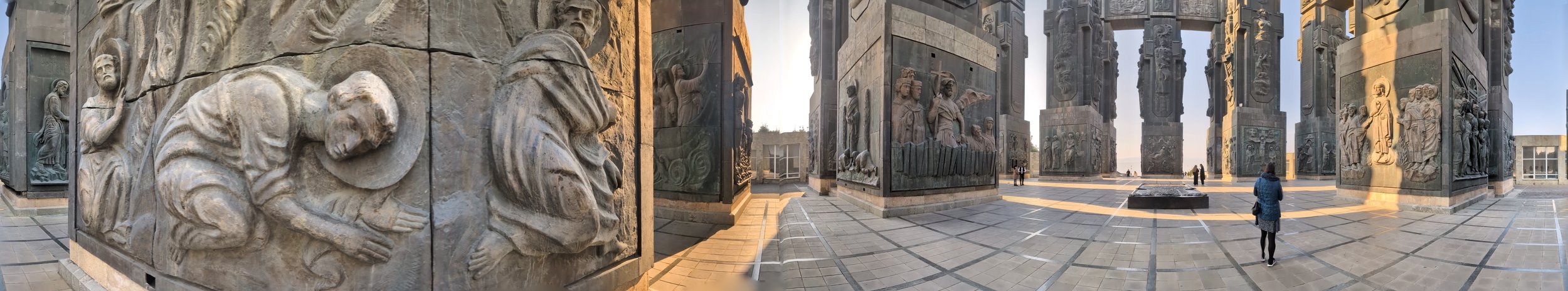 Panoramic view of the Motherland Calls monument with large stone relief sculptures on the base, depicting soldiers and historical scenes, under a clear sky, with a few people walking around in the plaza.