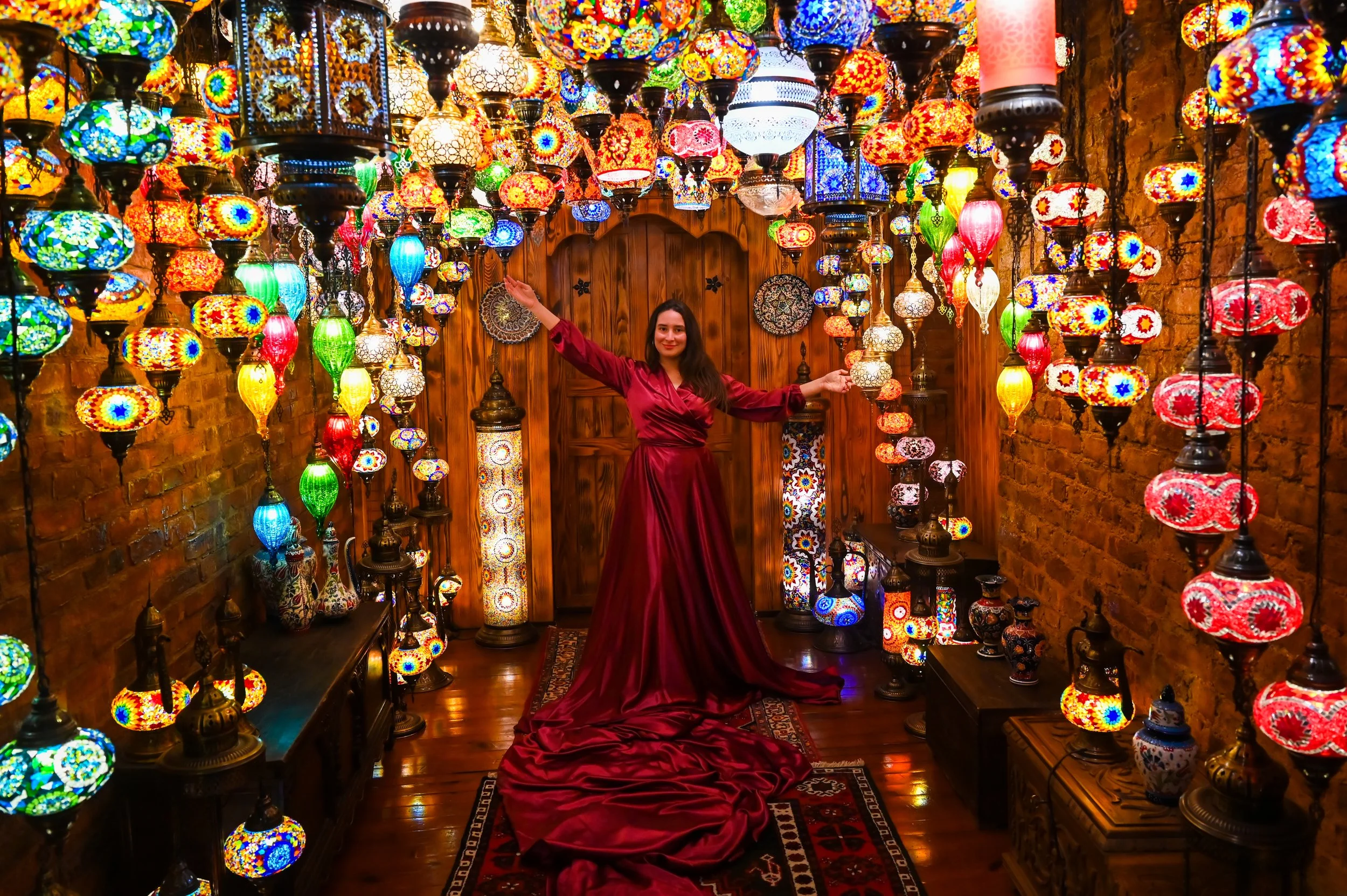 A woman in a long, red satin dress standing in a room filled with colorful Turkish lanterns hanging from the ceiling, with a wooden door and brick walls in the background.