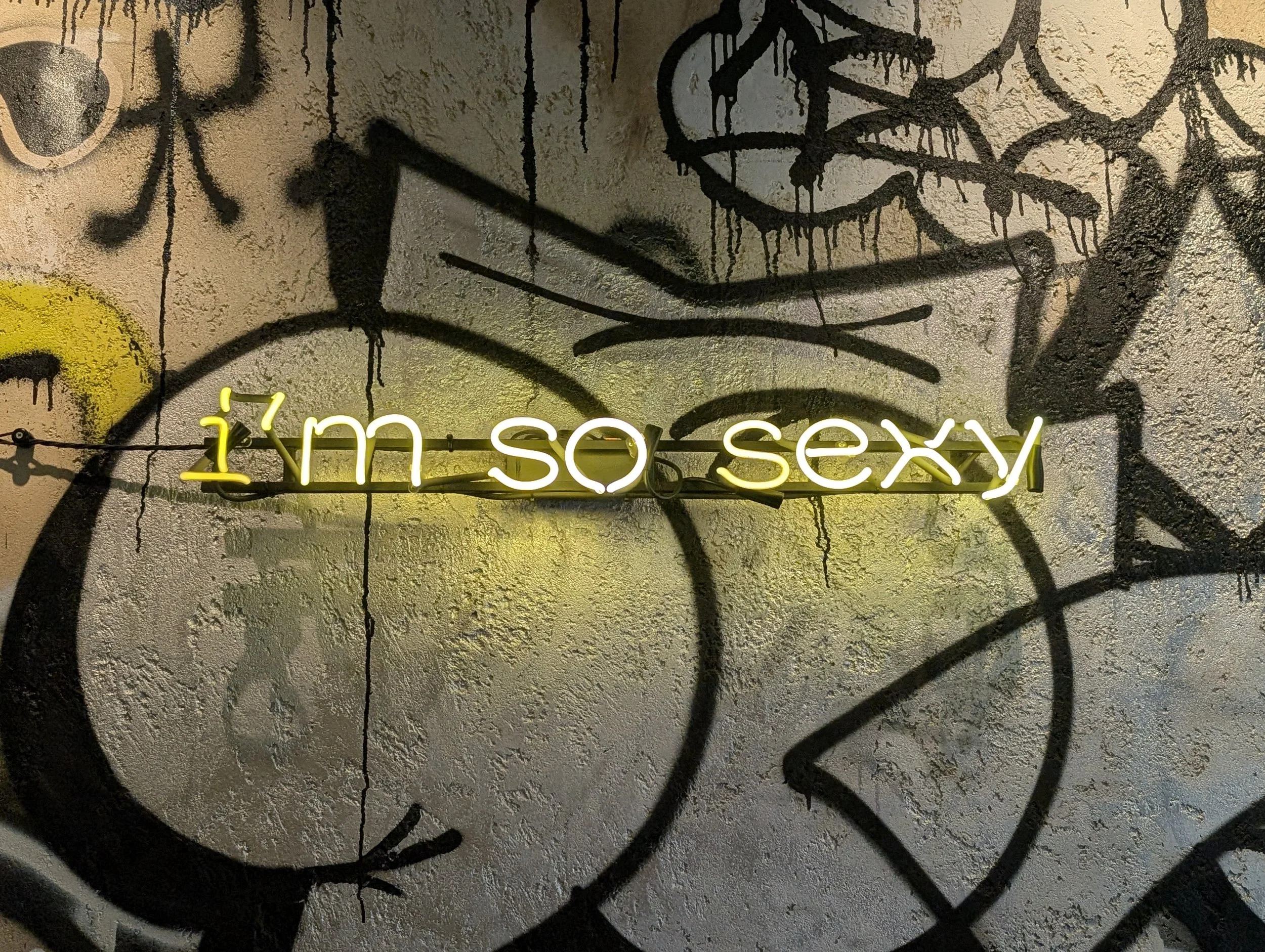 Graffiti art featuring black spray-painted abstract shapes and lines on a textured wall, with bright yellow neon sign reading 'it's so sexy' at the bottom.