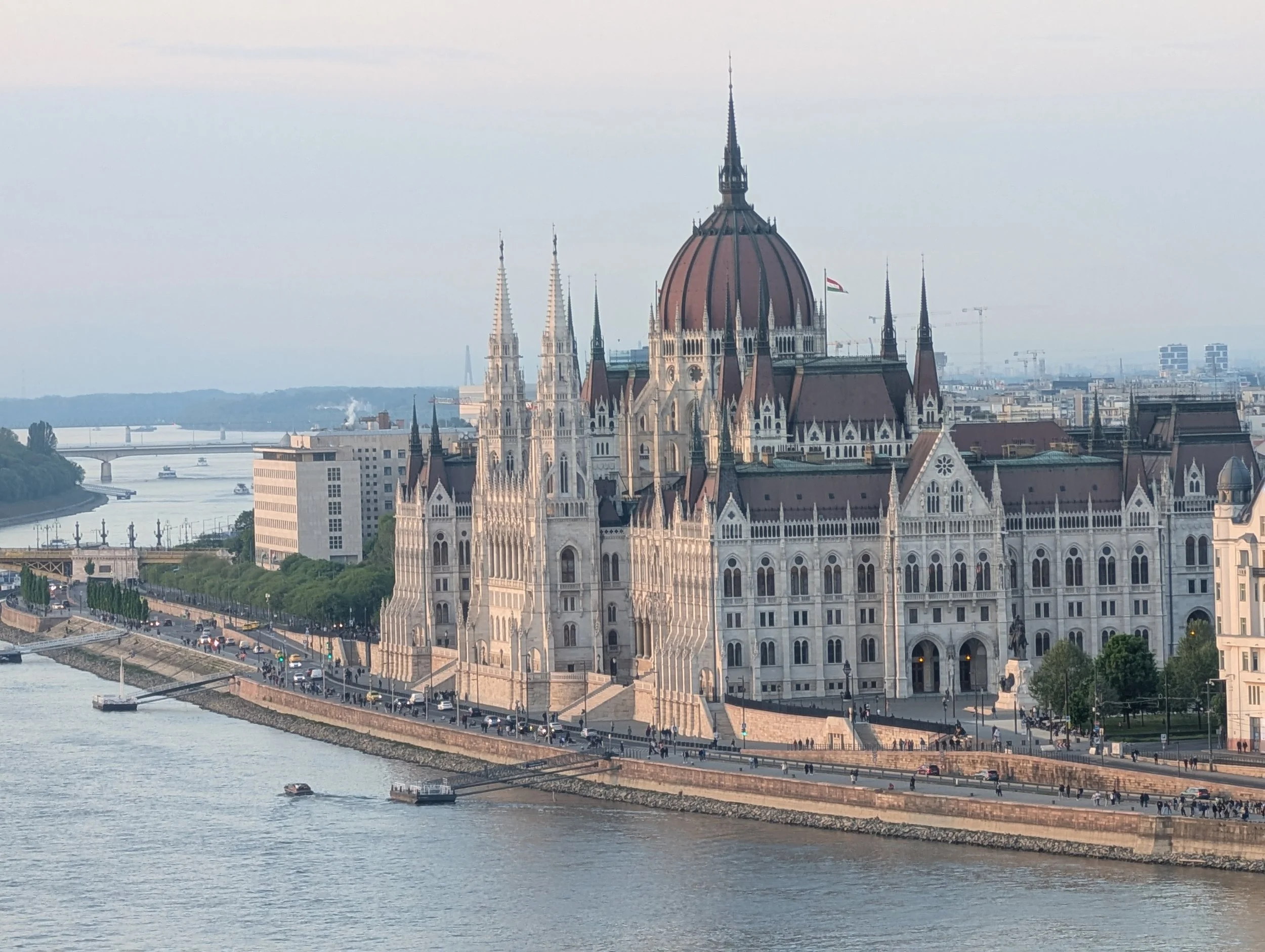 View of the Hungarian Parliament Building along the Danube River in Budapest, Hungary, with a boat on the water and a bridge in the background.