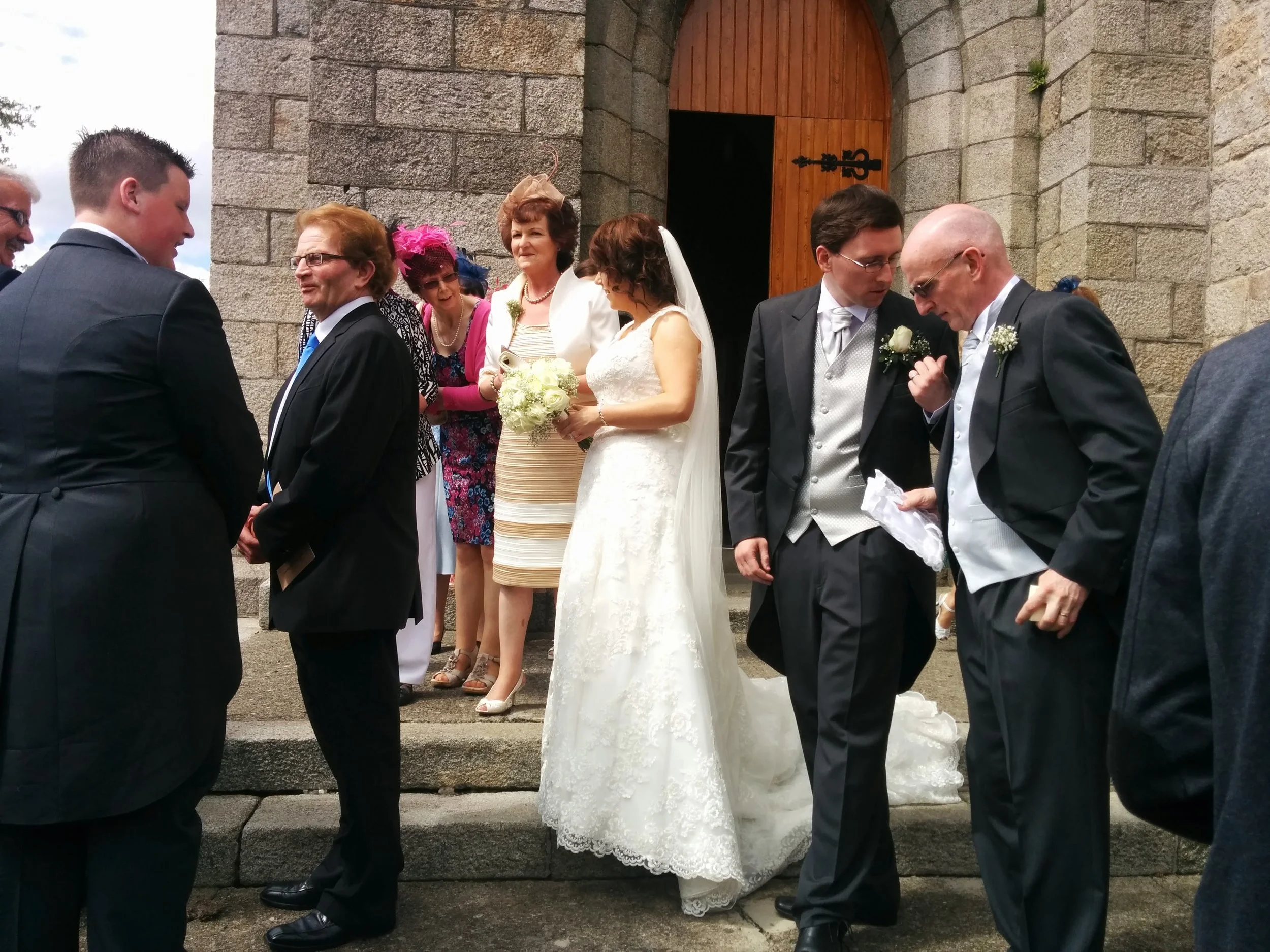 A group of wedding guests, including women in dresses and men in suits, standing outside a stone church with wooden door and black latch, participating in a wedding ceremony.