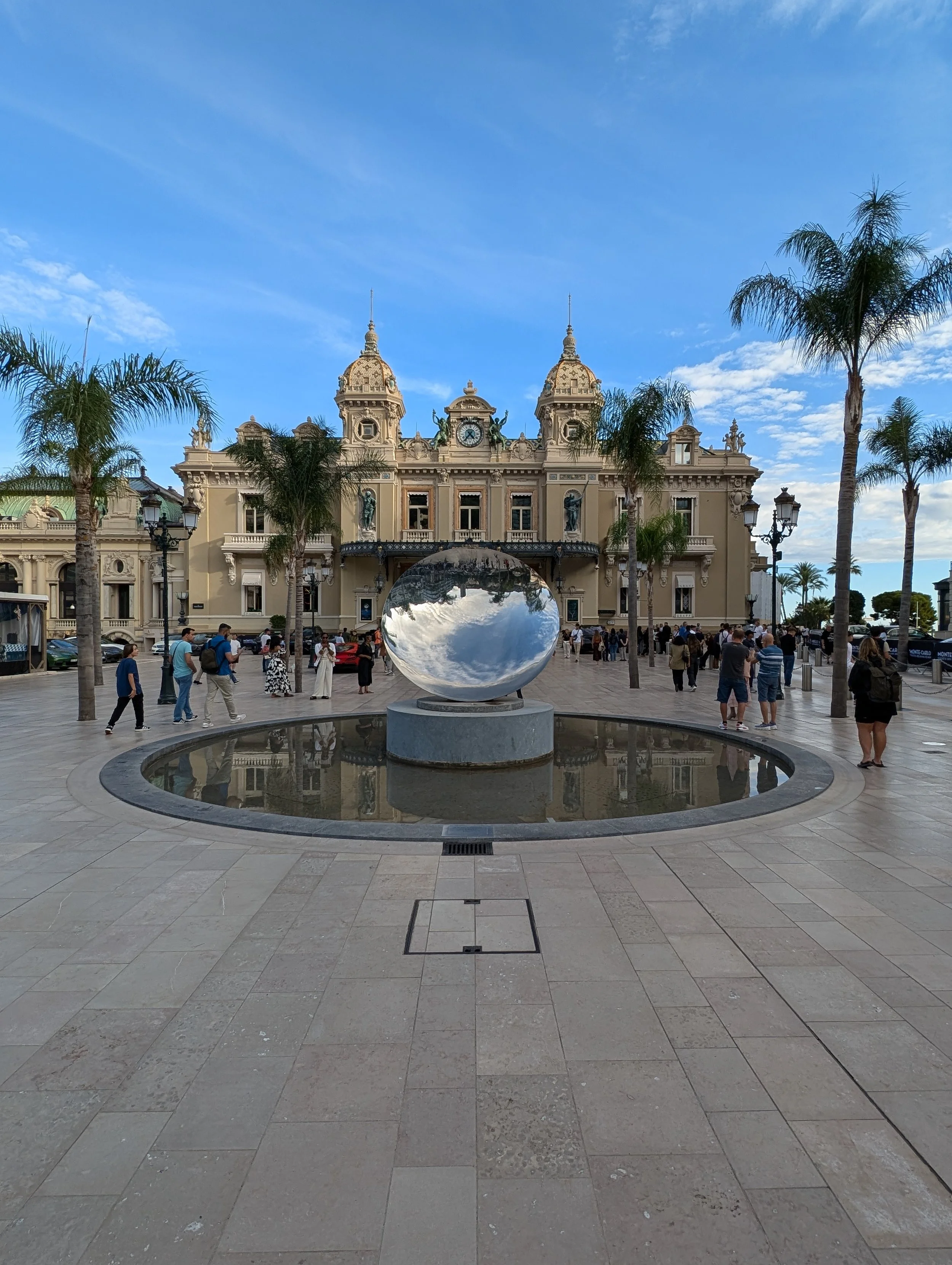 A historic building with ornate architecture, clock, and towers, surrounded by palm trees and a crowd of people in a town square, with a large reflective sculpture in the foreground.
