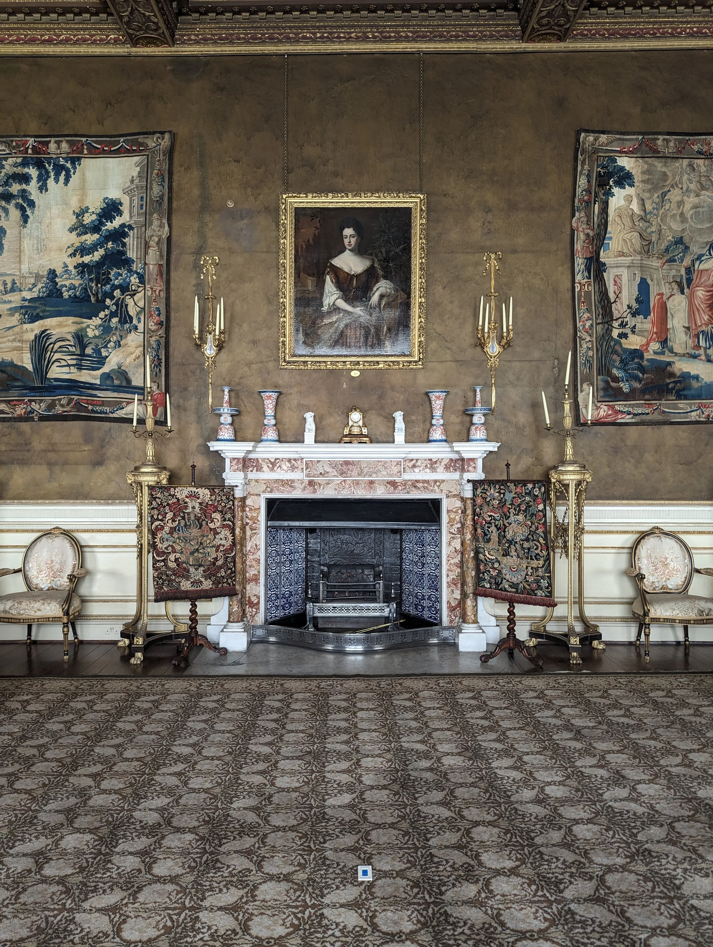 A decorated room with a large fireplace, ornate rugs, two chairs, a portrait of a woman above the fireplace, and tapestries on the walls.