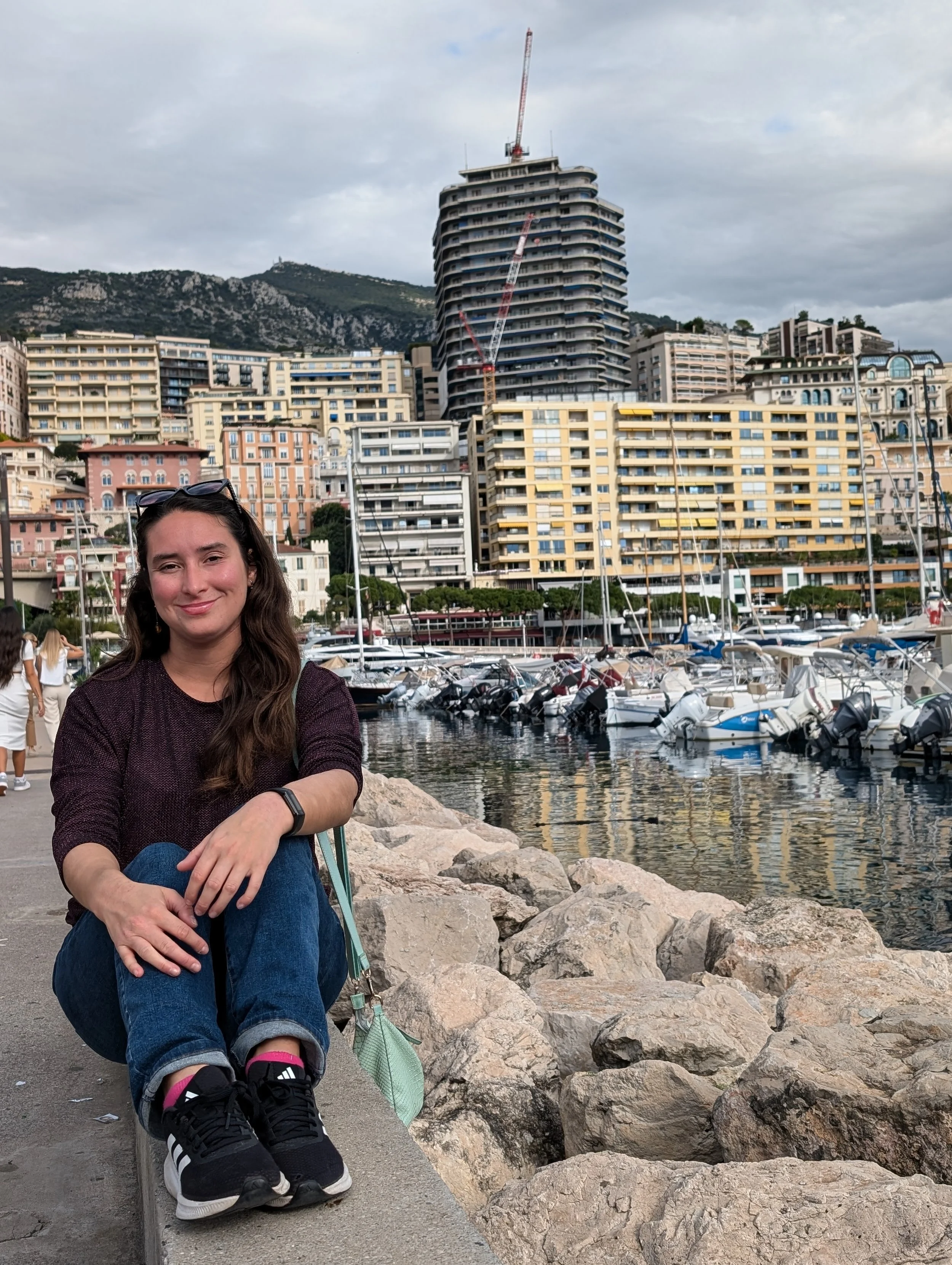 A woman with long dark hair sitting on a rock by the marina, with boats docked behind her and a cityscape with tall buildings and mountains in the background.