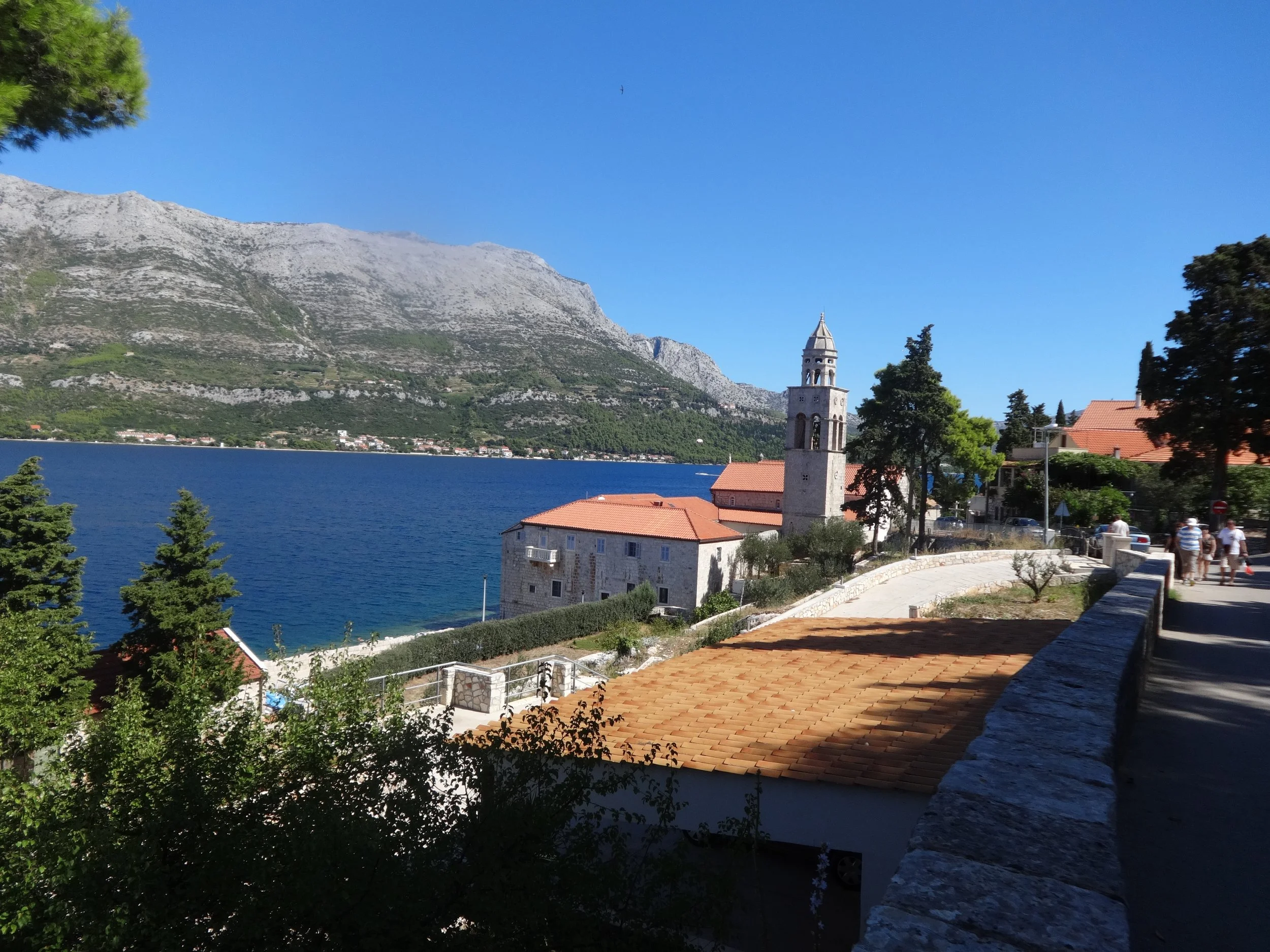A scenic view of a large lake with a historic church and red-roofed buildings along the shore, surrounded by green trees and mountains under a clear blue sky.