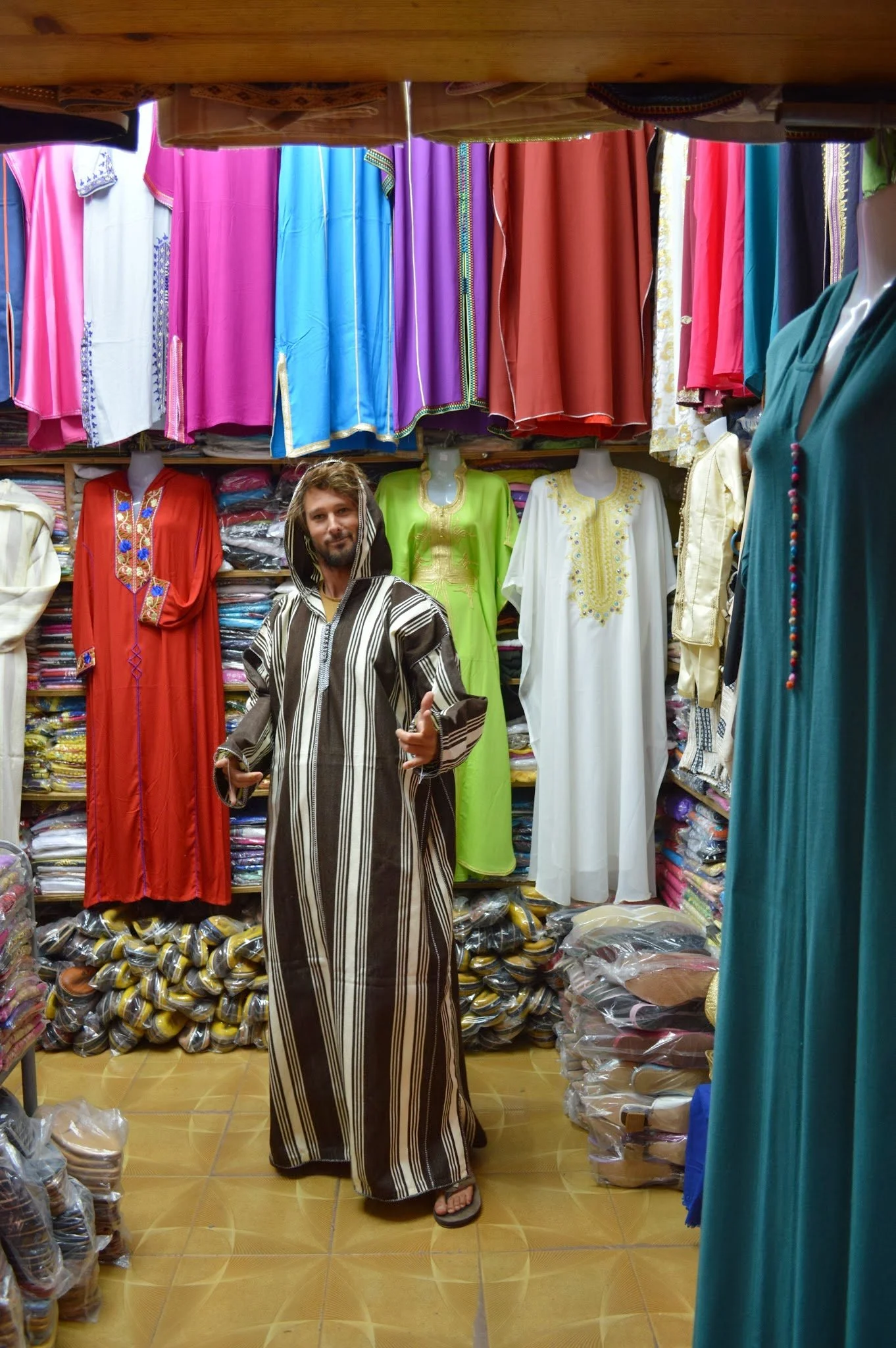A man with dreadlocks wearing a long, striped, traditional garment standing in a clothing store with colorful dresses and folded fabrics around him.