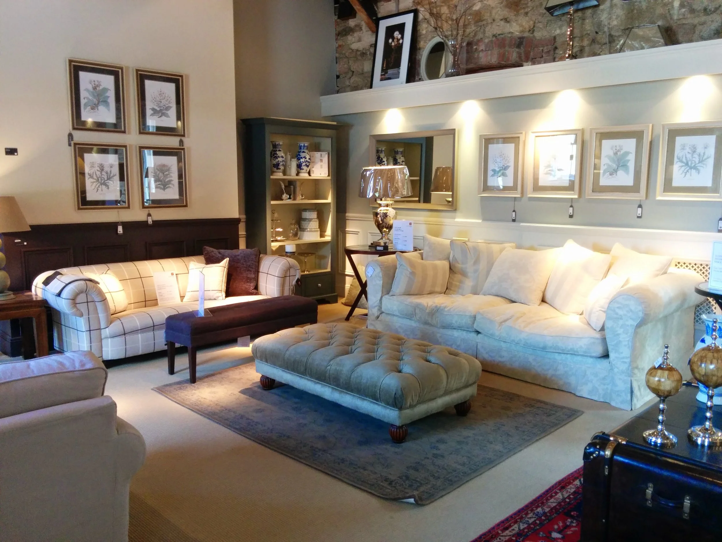 Interior of a furniture store displaying sofas, accent chairs, a rug, and decorative items with framed art on the walls, exposed brick and a gallery shelf.