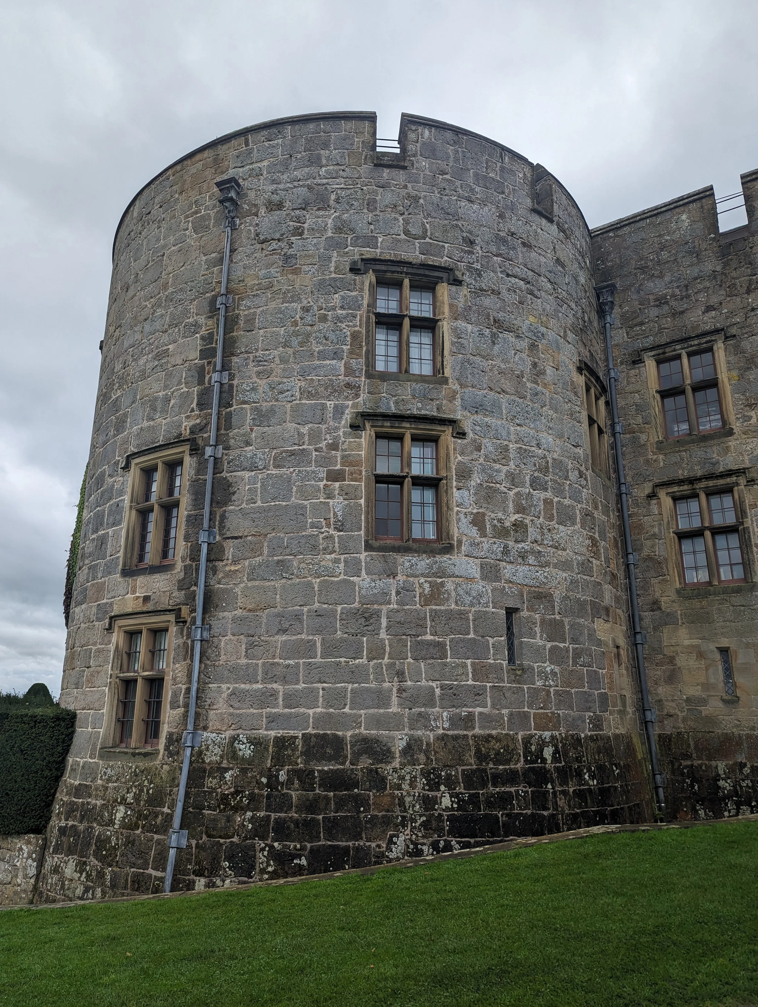 A stone castle with narrow windows and metal pipes on its exterior, surrounded by a grassy lawn and overcast sky.