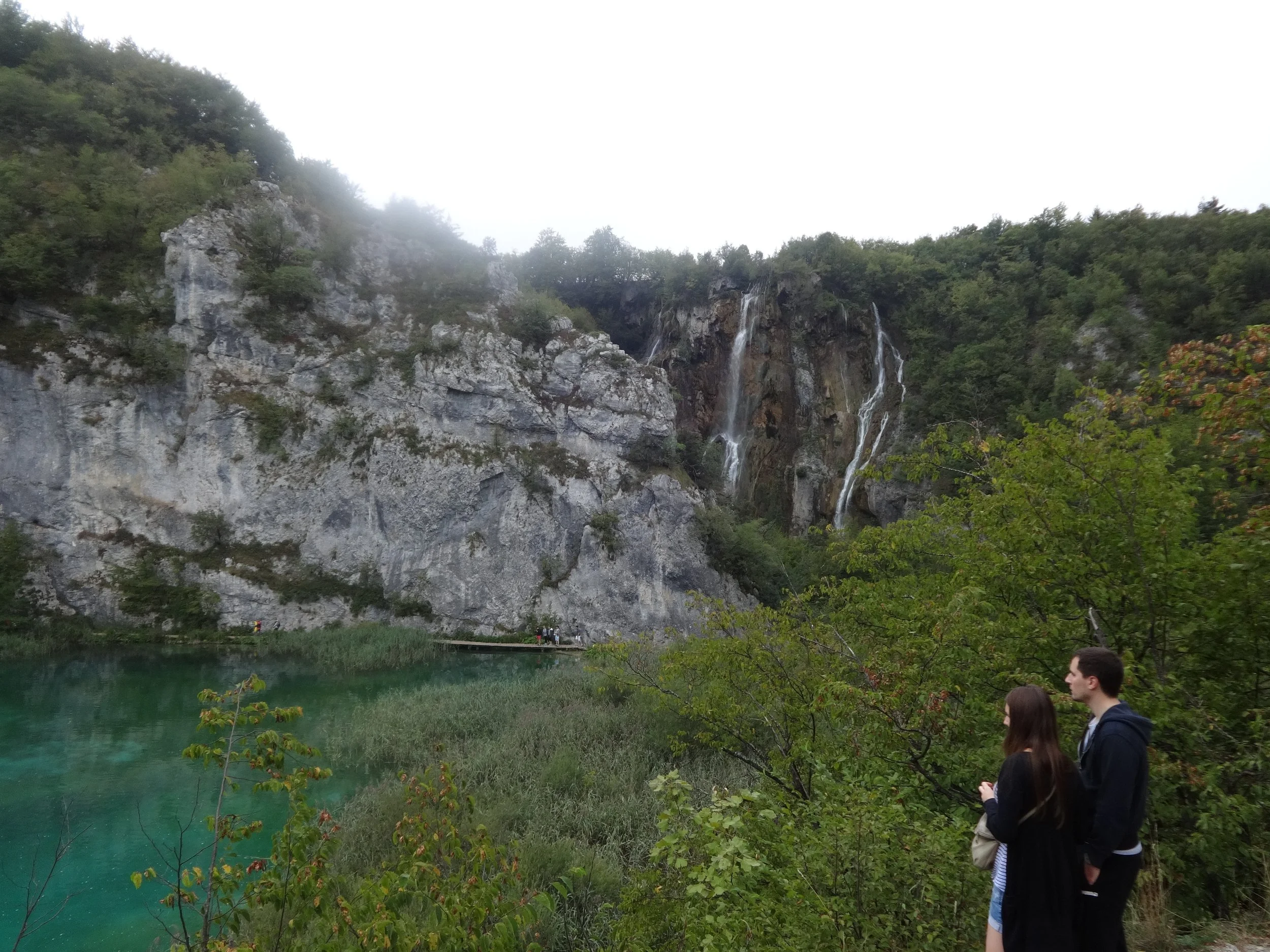 People stand near a turquoise lake at the base of a rocky mountainside with waterfalls and lush green trees.