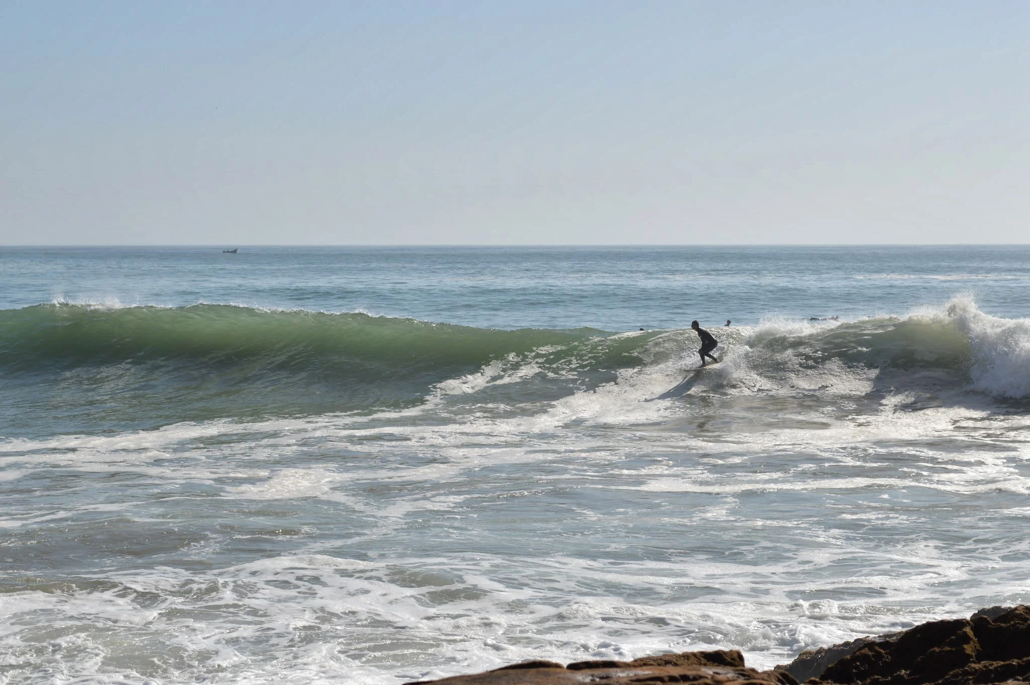A person surfing on a wave at the beach with a boat in the distance and a clear sky.