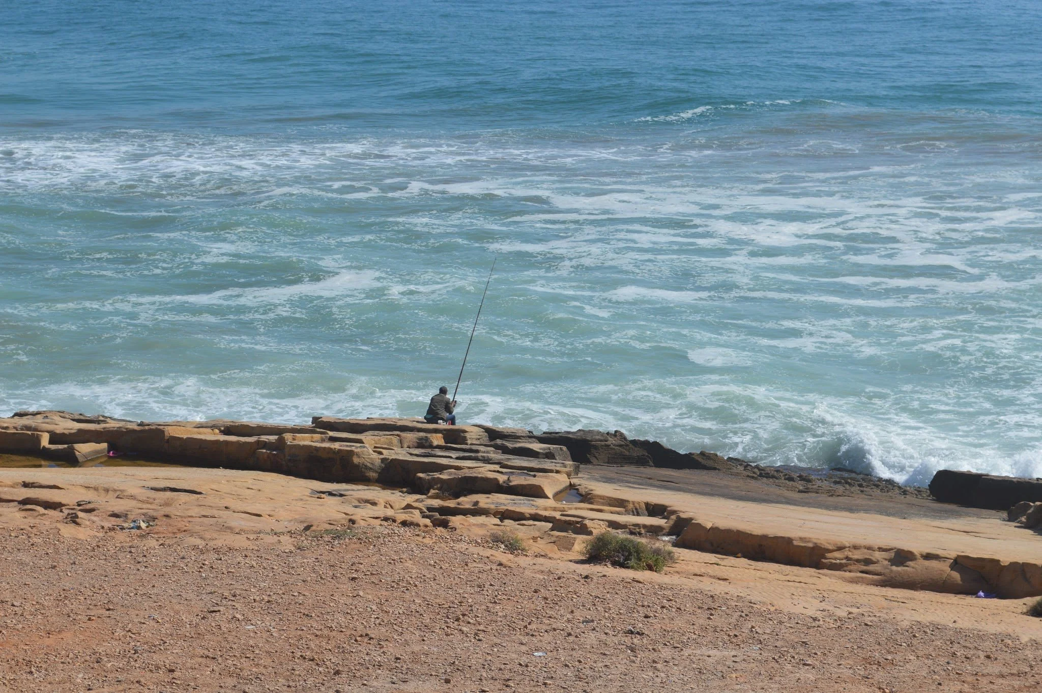 A person sitting on rocks by the shore, fishing in the ocean with waves and a blue sky in the background.