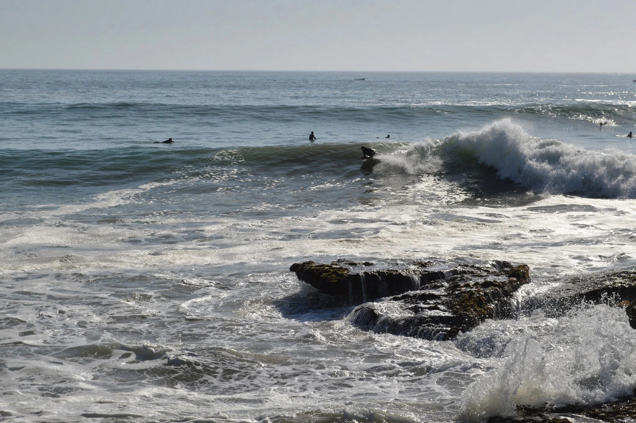 People surfing and swimming in the ocean with waves breaking near a rocky shoreline, and a boat visible in the distance.