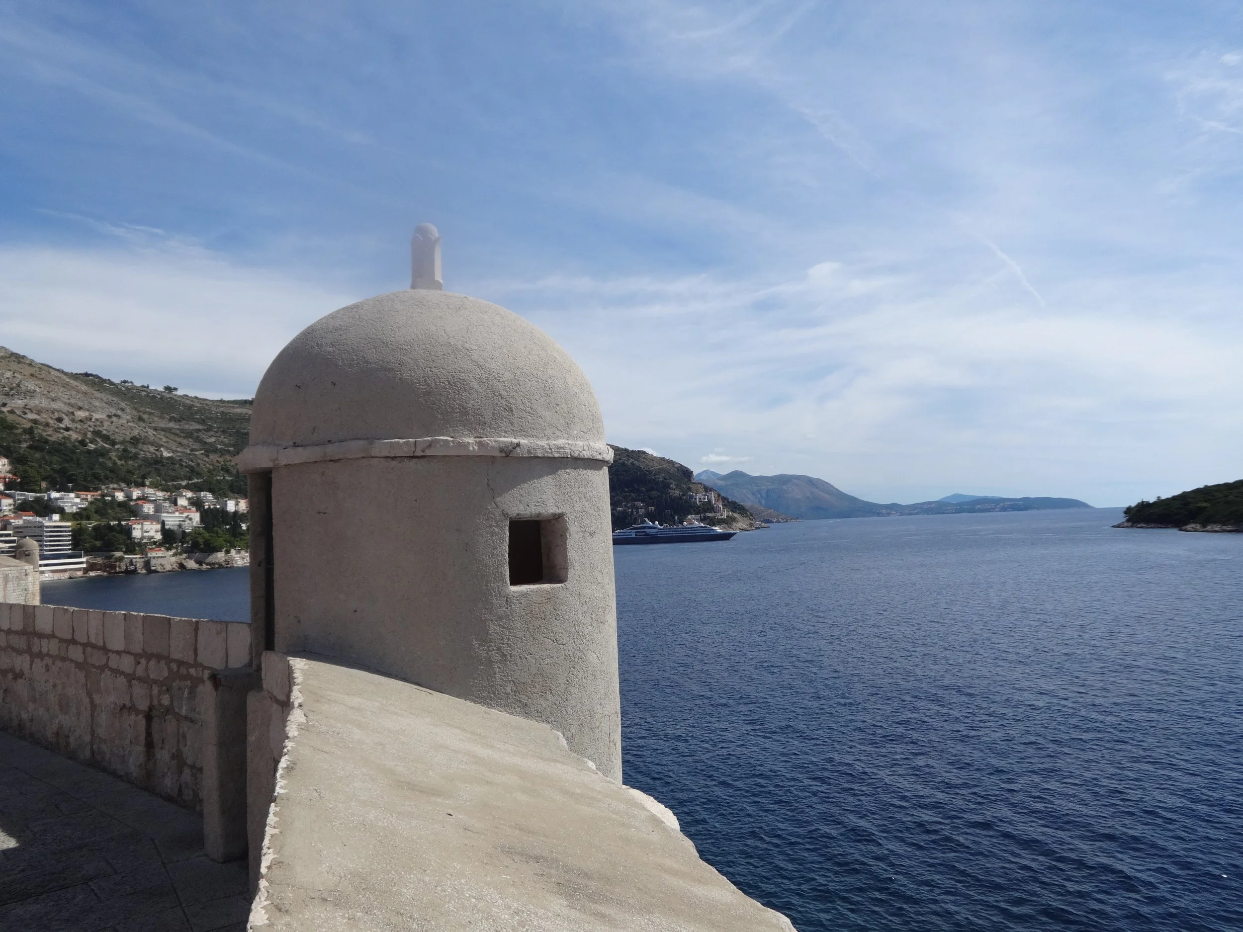 A white, rounded lighthouse with small window, situated on a coastal walkway overlooking water, with a hillside town nearby and mountains in the distance under a partly cloudy sky.
