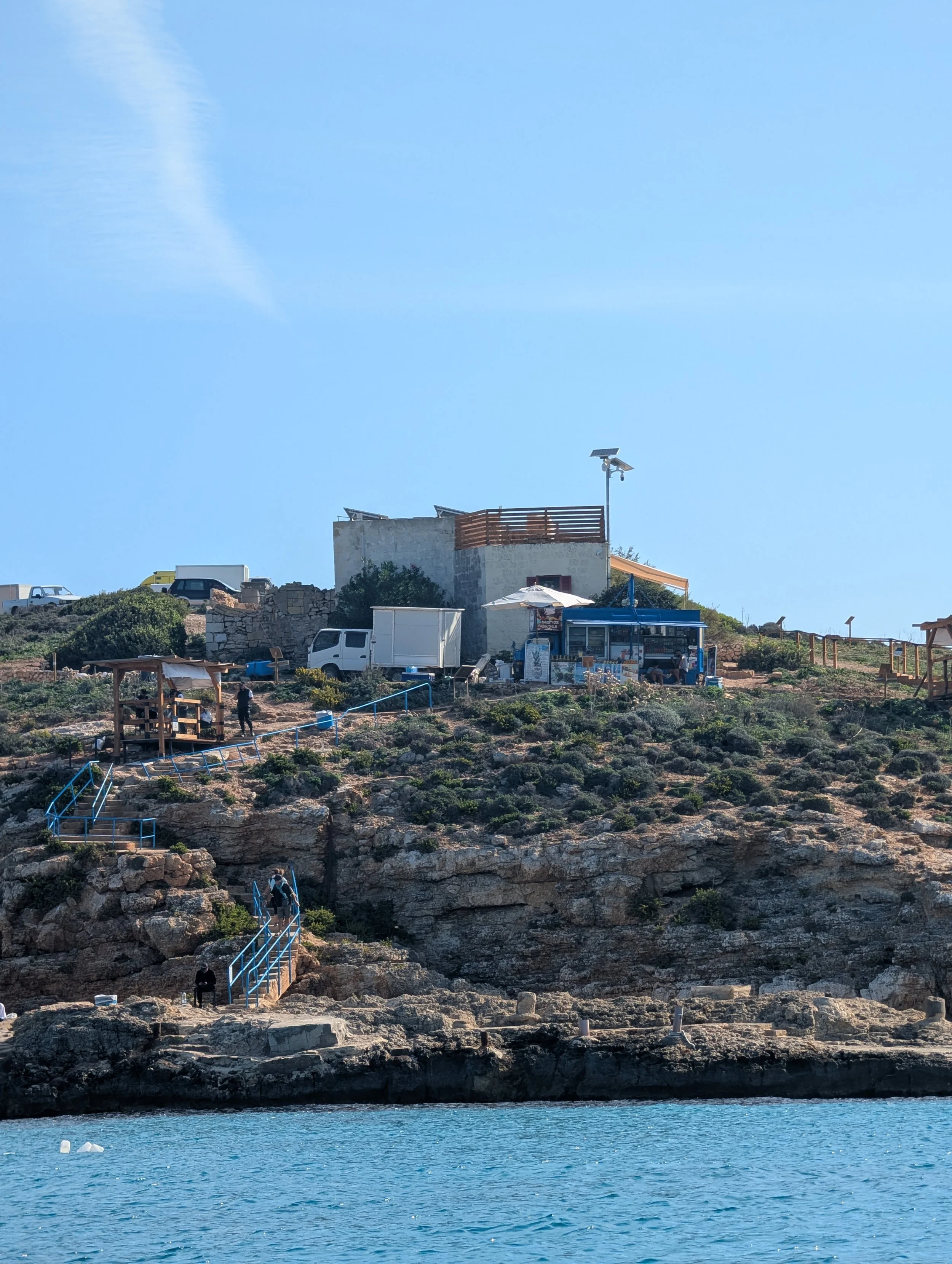 A hillside with stairs and pathways, a building with solar panels, and a body of water in the foreground under a clear blue sky.