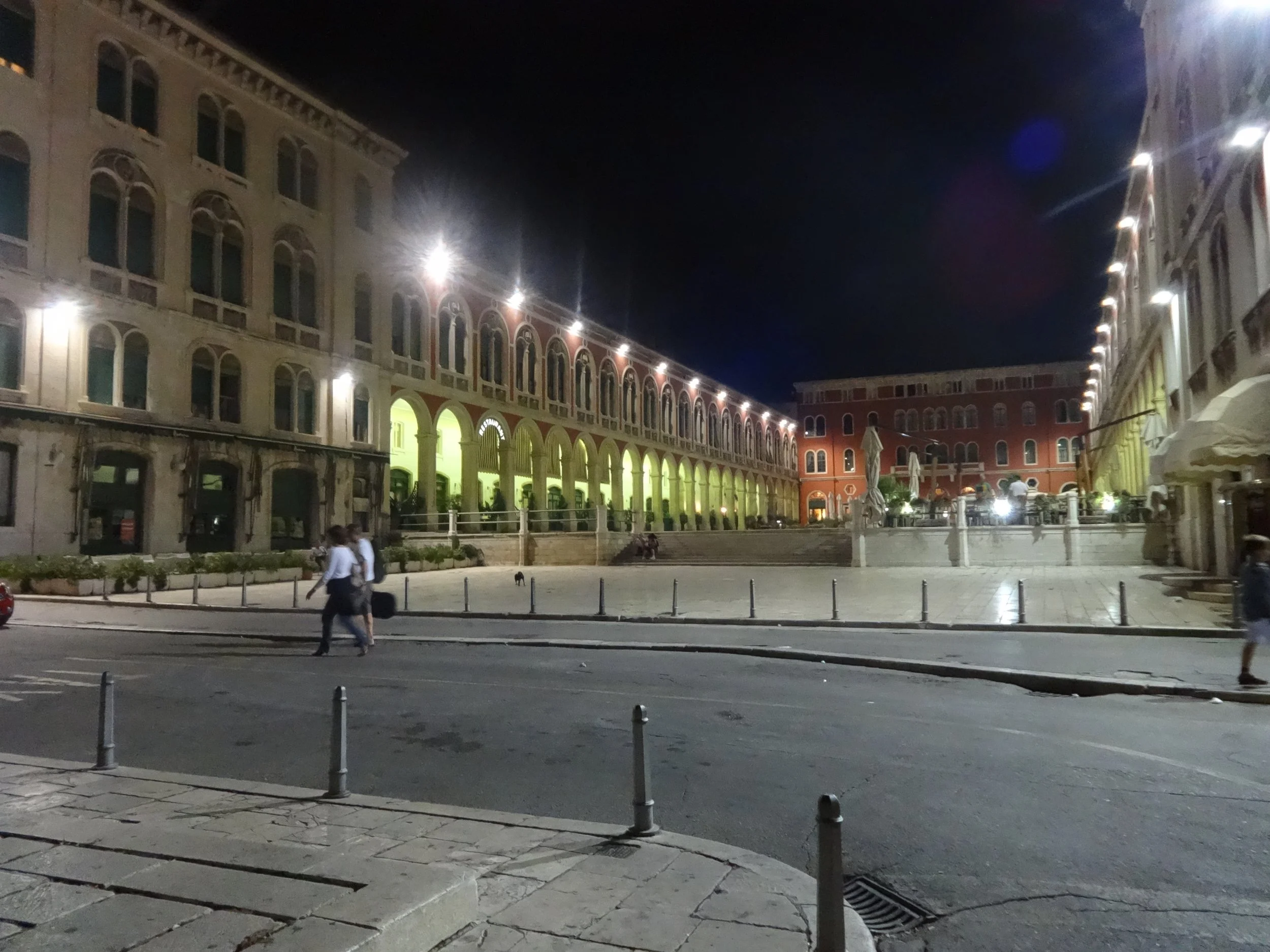 Nighttime scene of a city square with historic buildings, some illuminated, and a few people walking near the parked cars, with umbrellas and outdoor seating visible.
