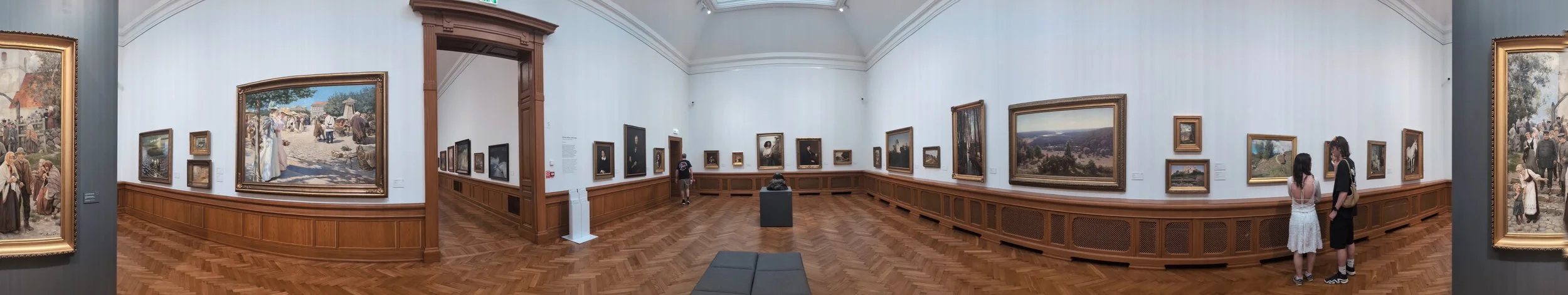 Panoramic view of an art gallery with white walls, hardwood floors, and framed paintings, with visitors observing the artwork.