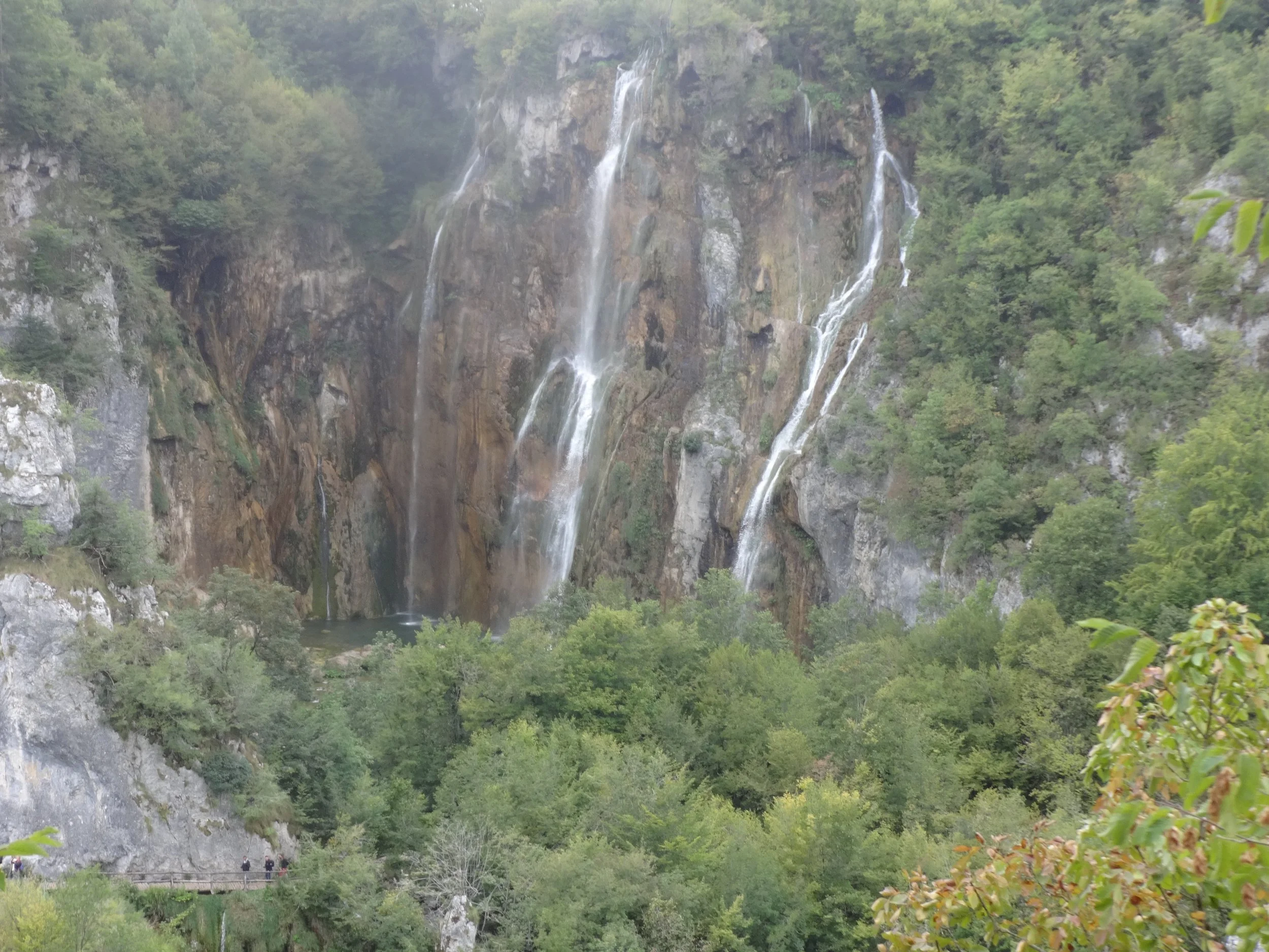 Waterfalls cascading down a rocky cliff surrounded by dense green forest, with a viewing platform and people at the bottom.