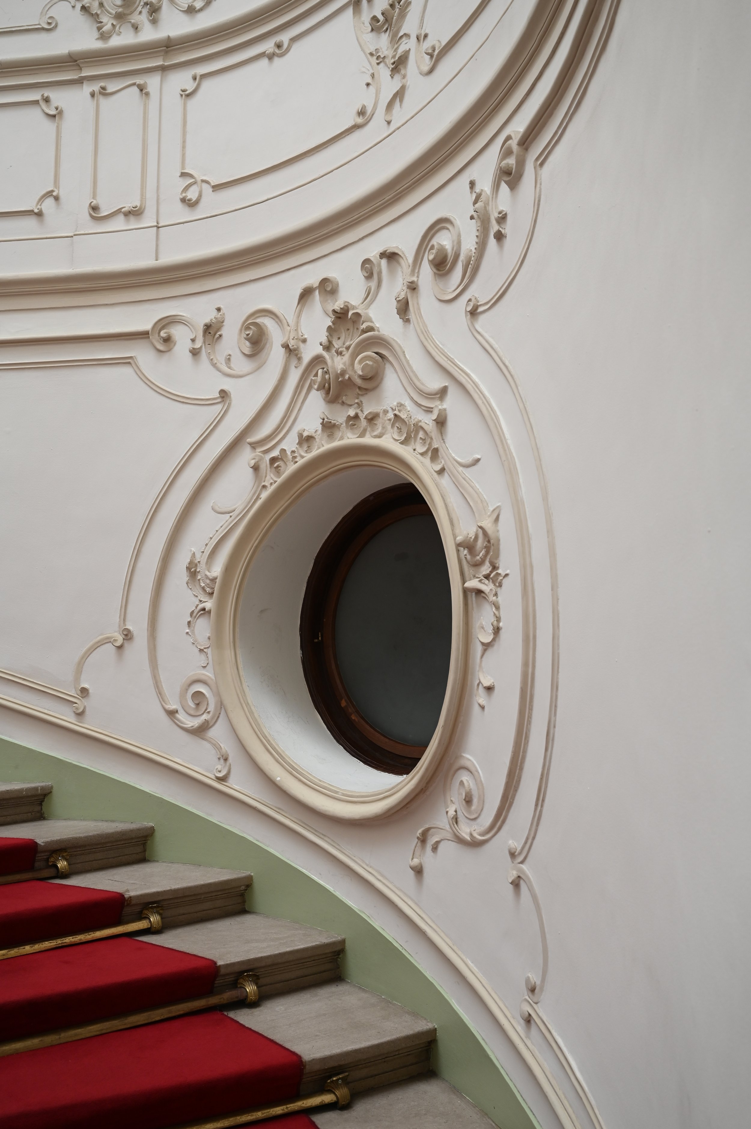 A curved staircase with beige steps, red carpet runners, and gold-colored railings. The wall beside the staircase features ornate, decorative plasterwork with intricate scrolls and floral motifs surrounding a circular window.