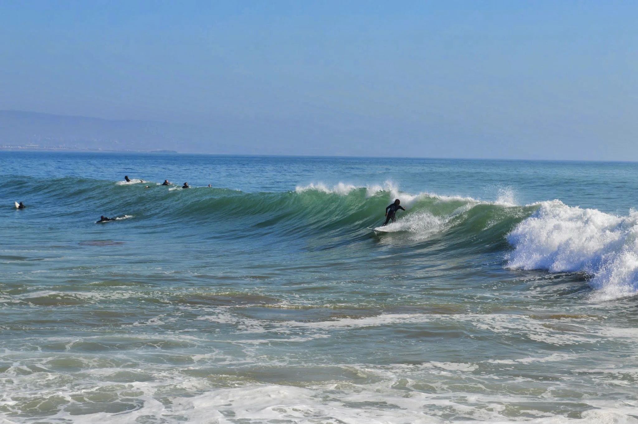 A person surfing on a wave in the ocean with several other surfers in the background and a distant landmass under a clear blue sky.