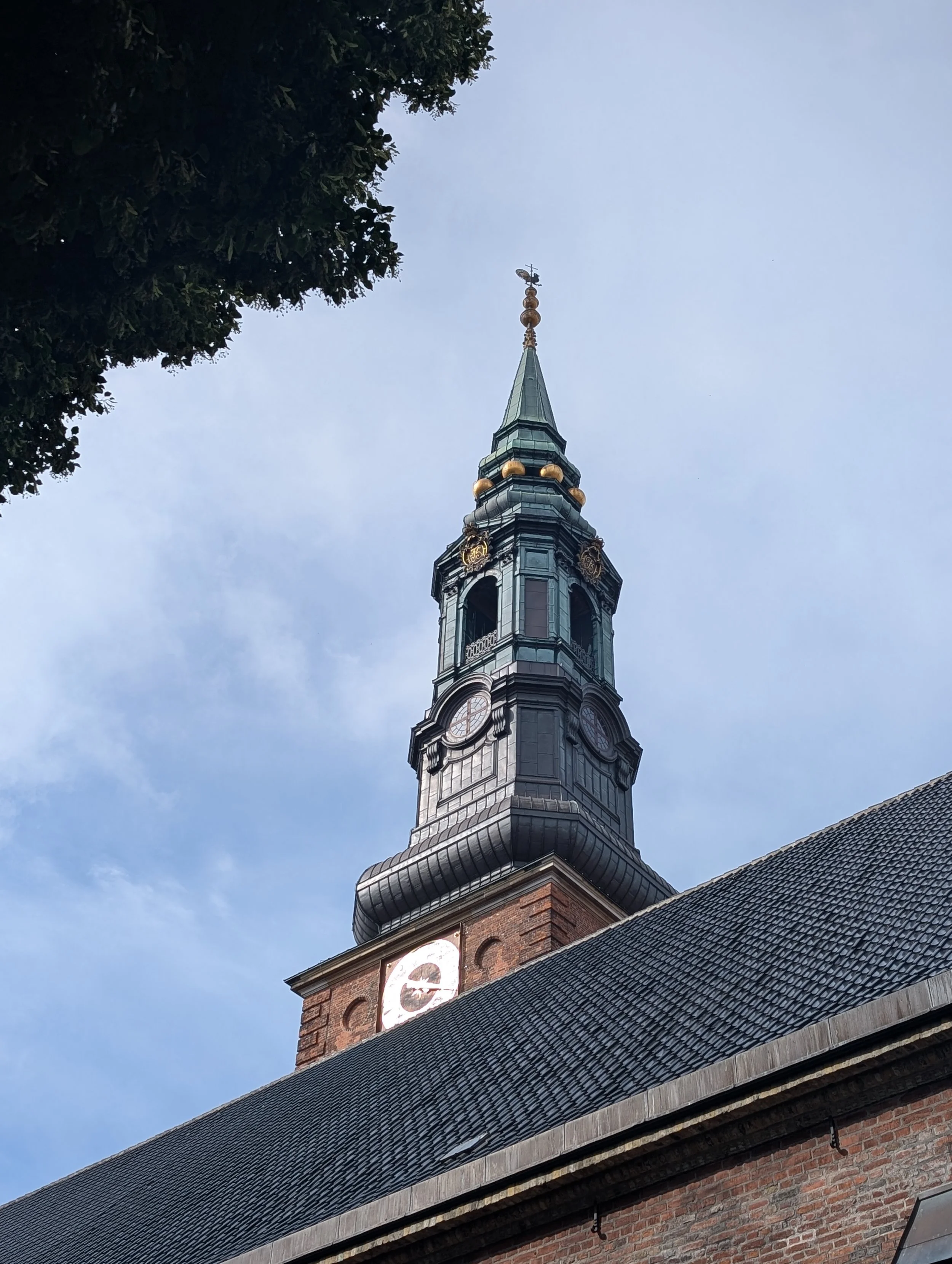 A historic brick church tower with a clock and a steeple topped by a cross, partially obscured by a tree, under a blue sky.