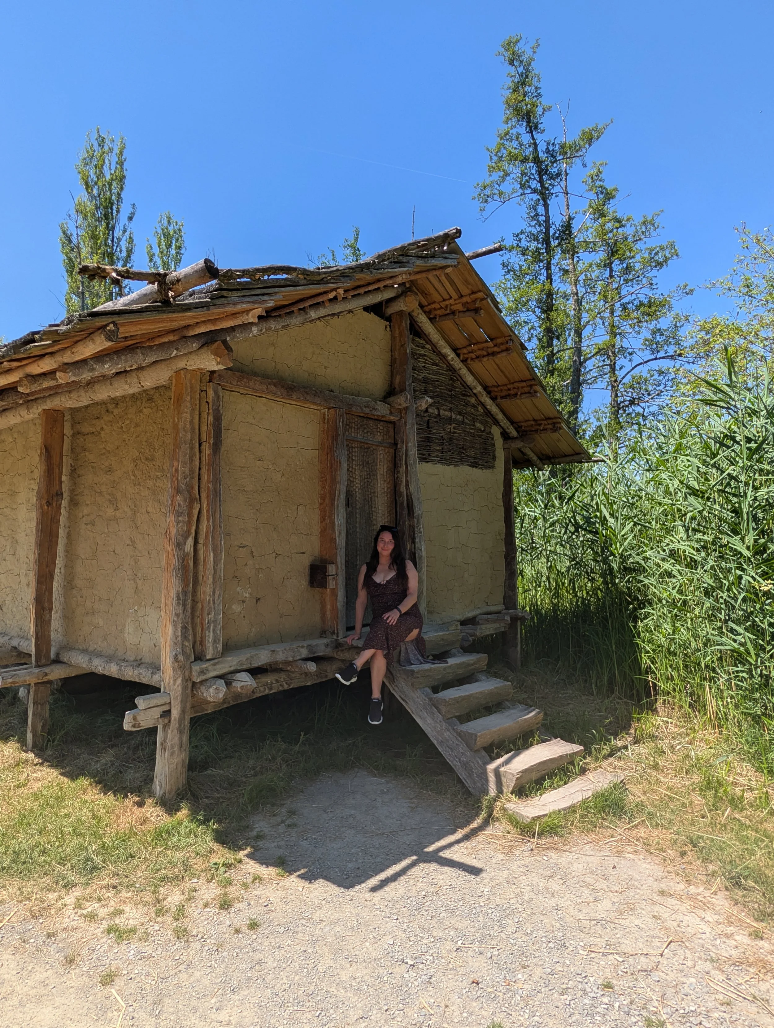 A woman sitting on wooden stairs outside a small, rustic house made of mud and wood, with a roof constructed of sticks and logs. The house is surrounded by tall green grass and trees under a clear blue sky.