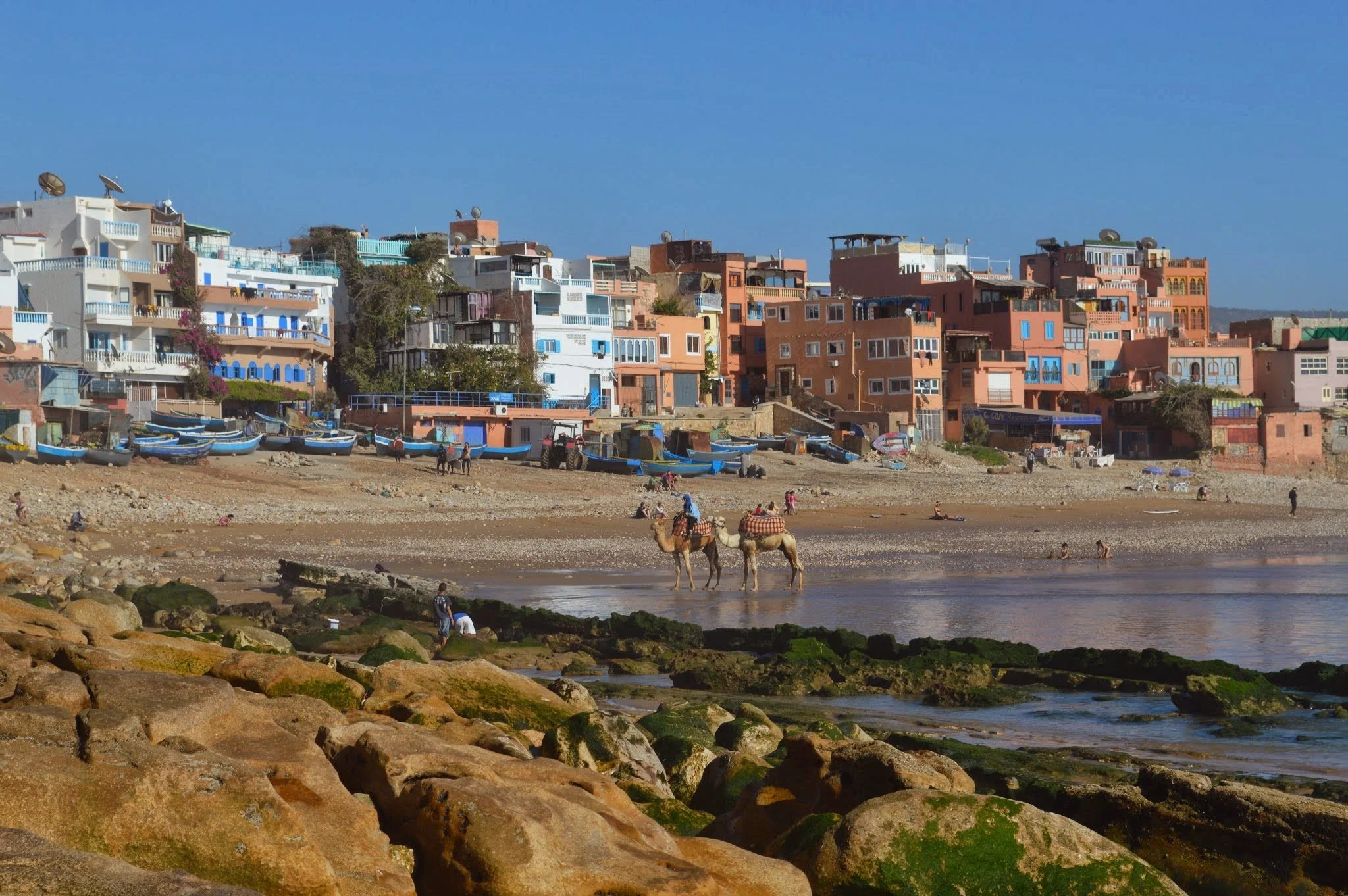 Colorful hillside buildings overlooking a beach with boats, people, and camels.