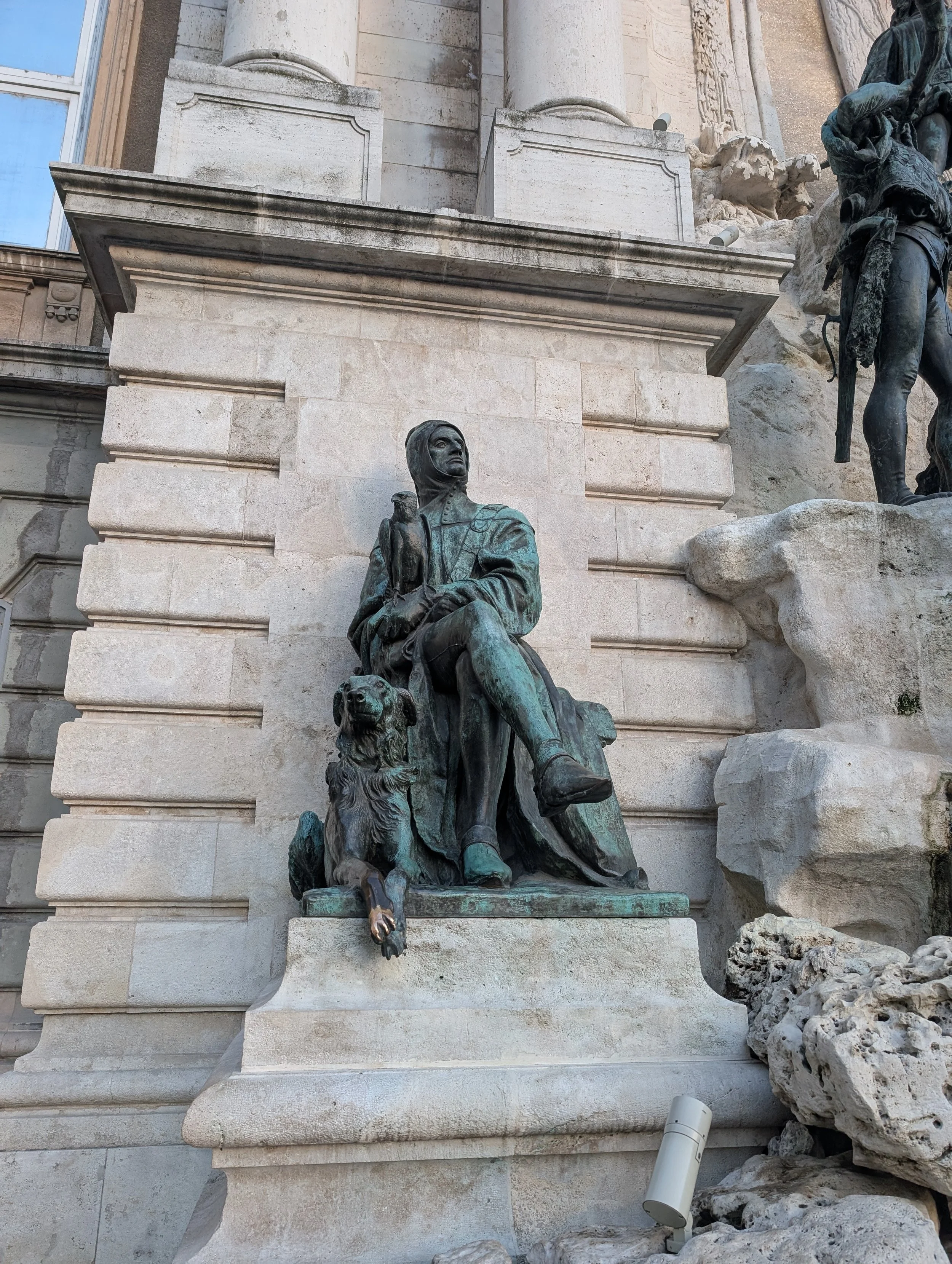 Statue of a seated man with a dog at his side, positioned in front of a large stone building with columns and decorative architectural features.