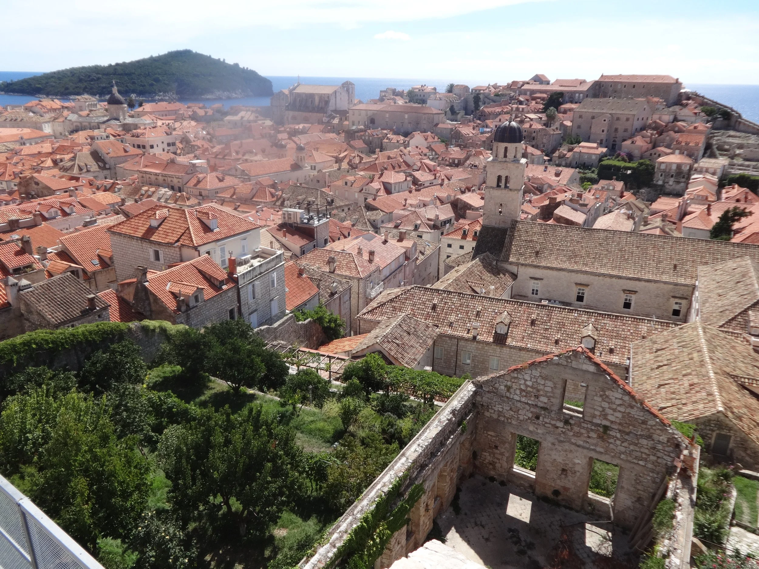 Photo of an old Mediterranean town with a dense cluster of historic stone buildings and red-tiled roofs, surrounded by greenery and overlooking the sea.