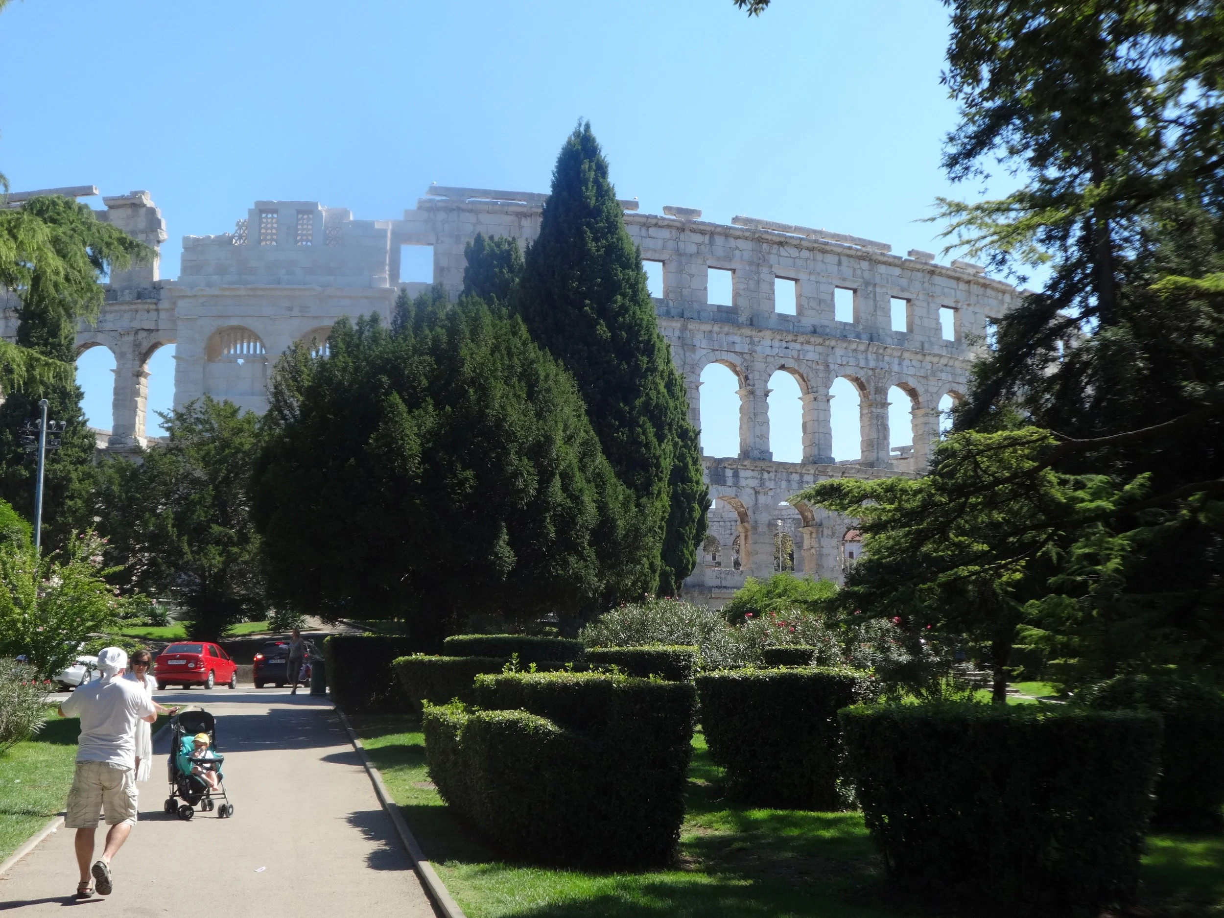 People walking in a park with the ancient Roman Colosseum in the background, partially obscured by trees.