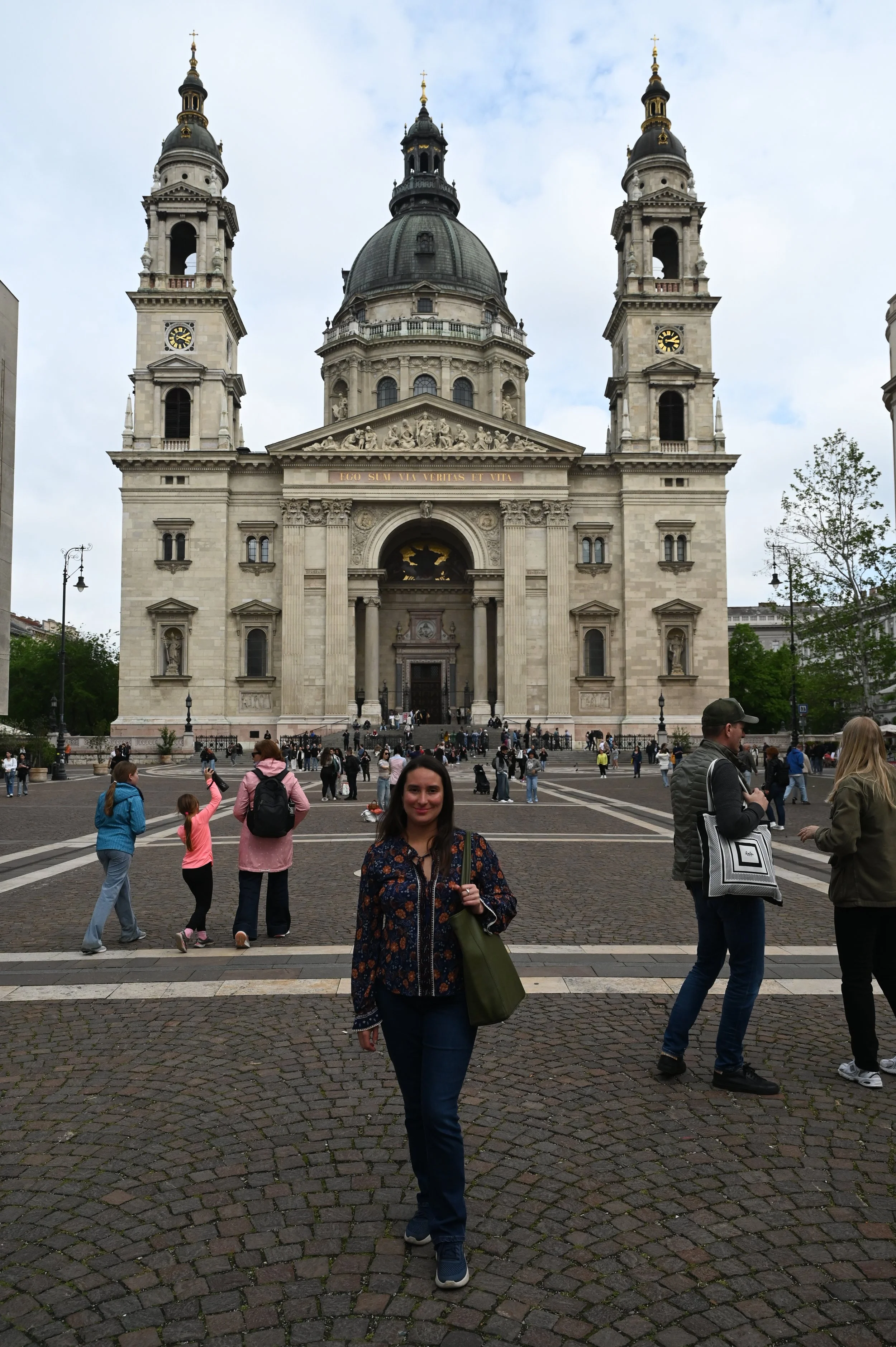 A woman walking on a cobblestone square in front of a large, historic church with a central dome and two bell towers. Several people are in the background near the church, some taking pictures and others walking.