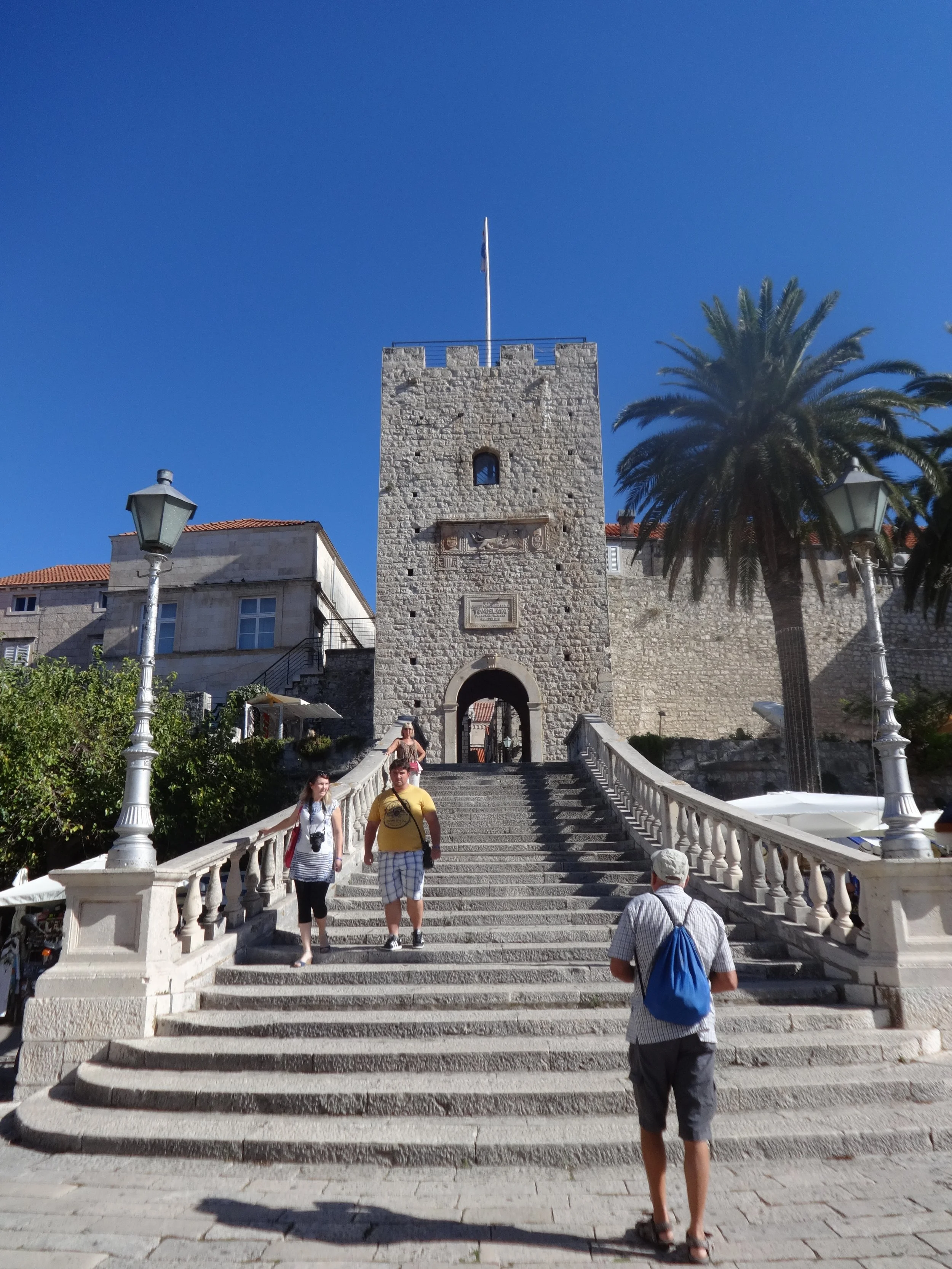 Stone tower with a flag on top, stairs leading up to it, and people walking on the stairs, with a palm tree and lamp posts nearby under a clear blue sky.