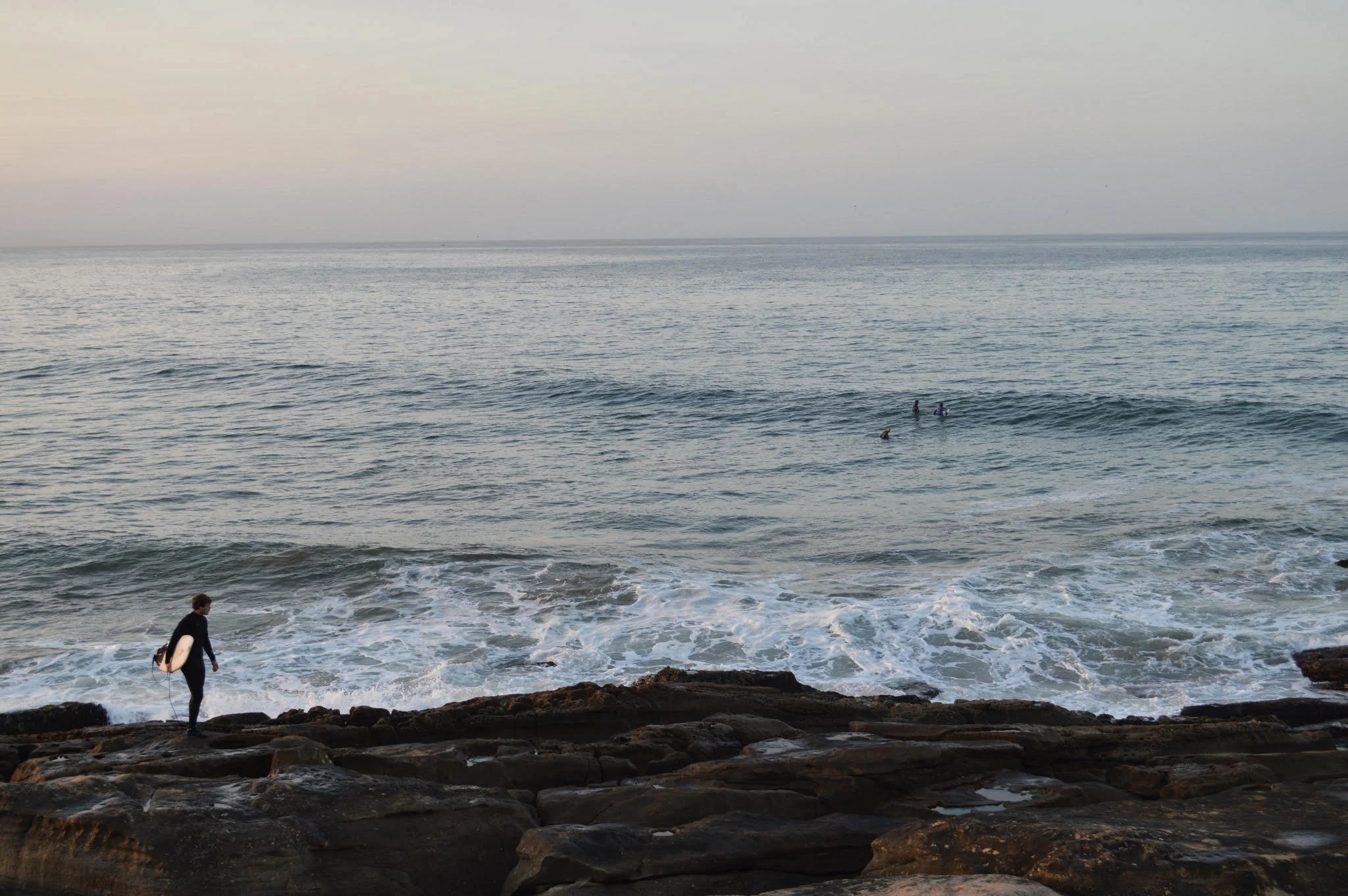 A person with a surfboard walking along rocky shoreline towards the ocean, with a few surfers in the water and a calm, slightly gray sky overhead.
