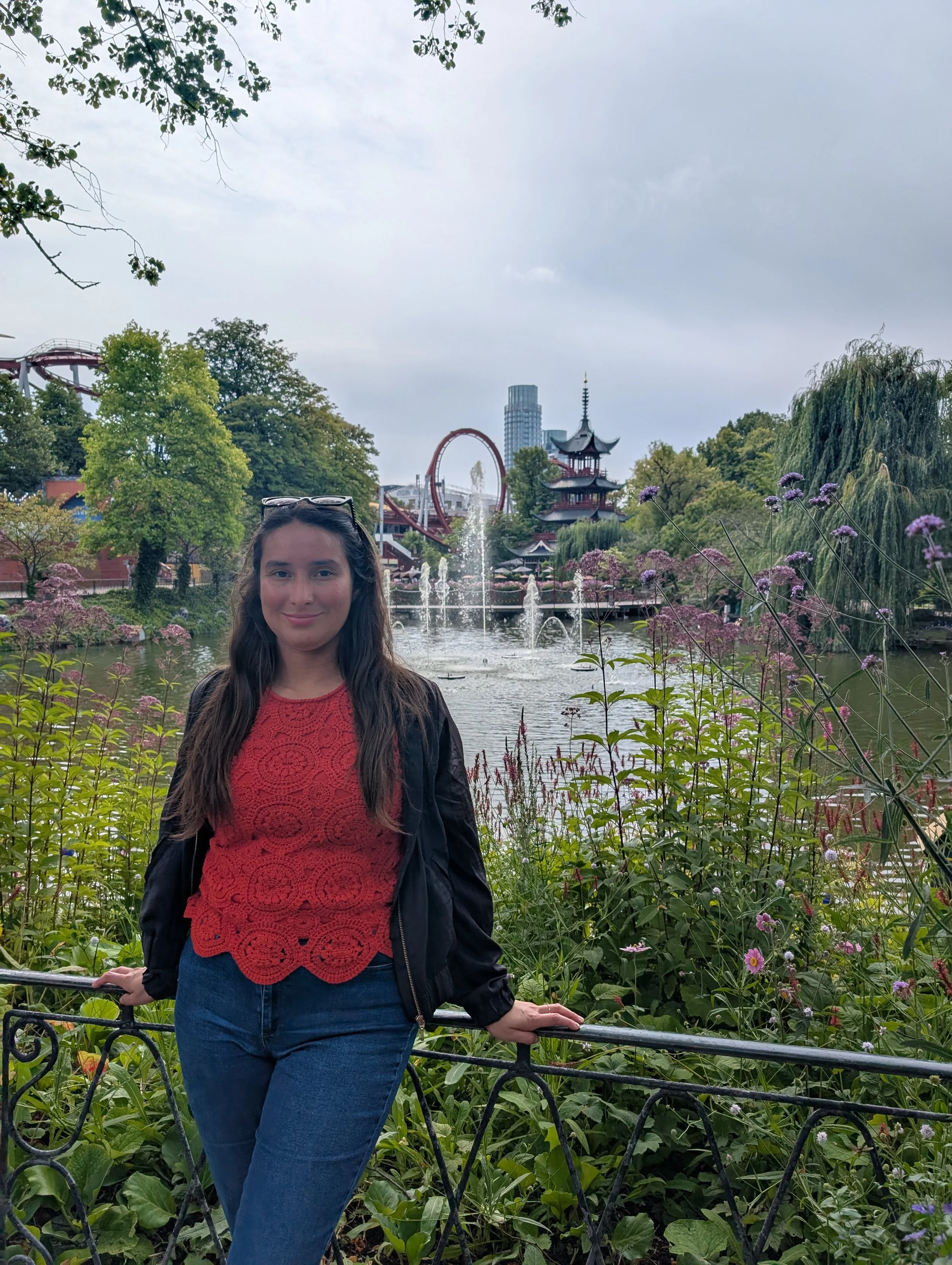 Young woman in red top and blue jeans standing outdoors in a park with water fountains and traditional Asian-style pagoda in the background, surrounded by green trees and purple flowers.