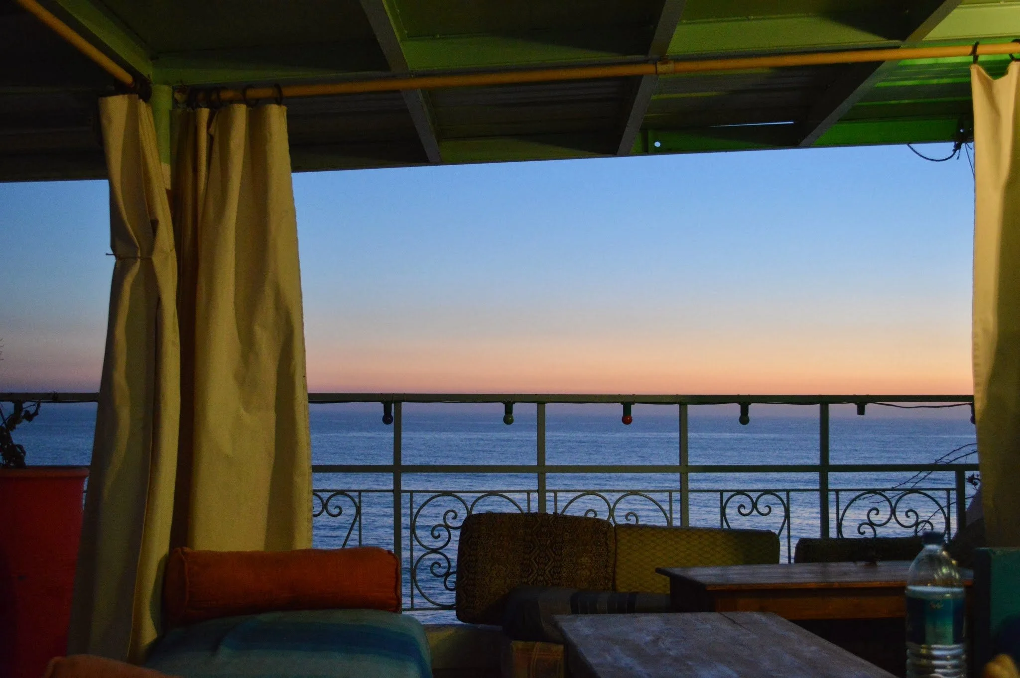 View of the ocean at sunset as seen from a balcony with curtains, chairs, and a table.