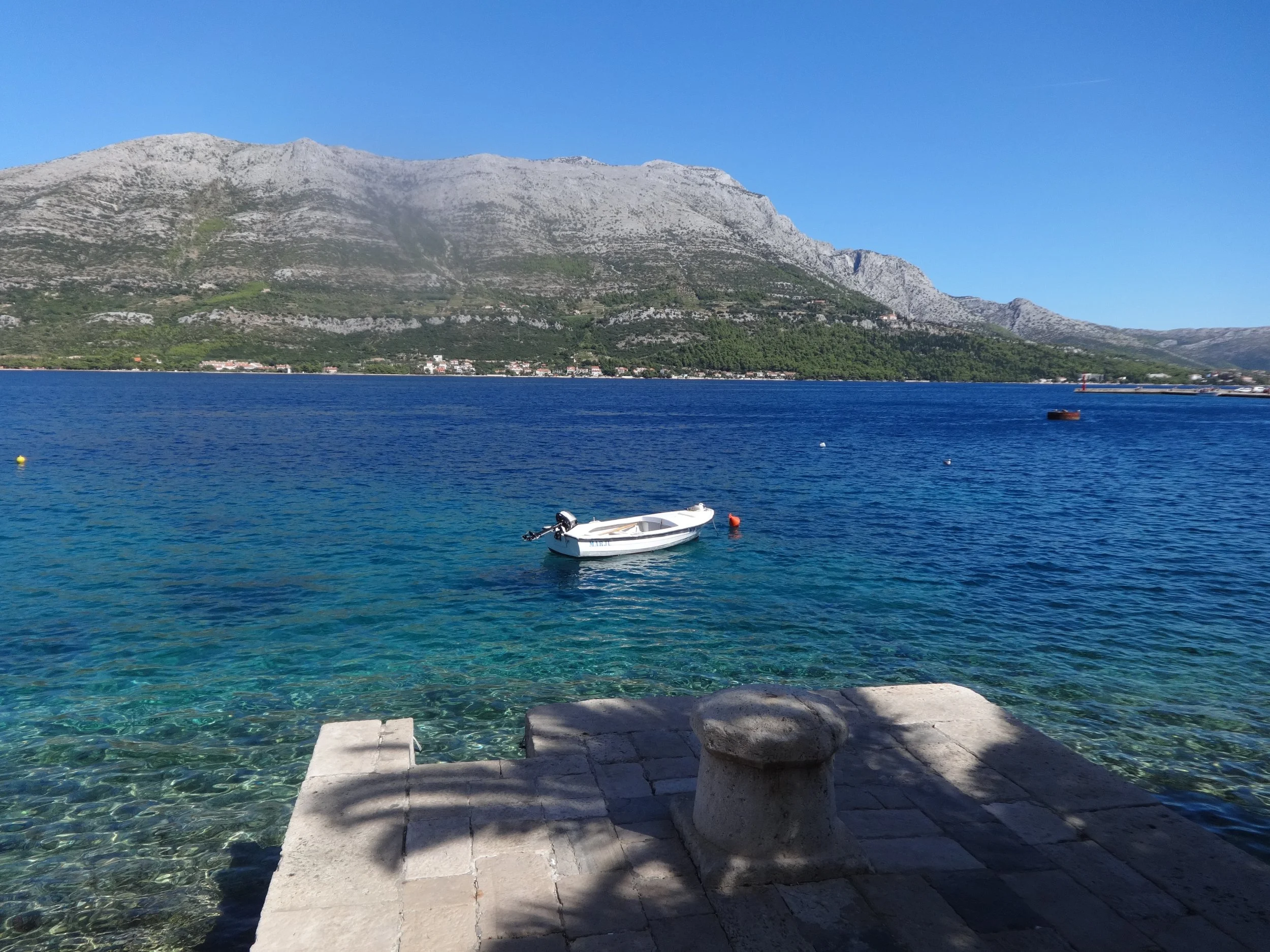 A scenic view of a mountain landscape with a calm blue body of water in the foreground. There is a small white boat floating on the water near the shore, and a rocky pier or platform in the foreground with stone benches. The mountains are grey and gr