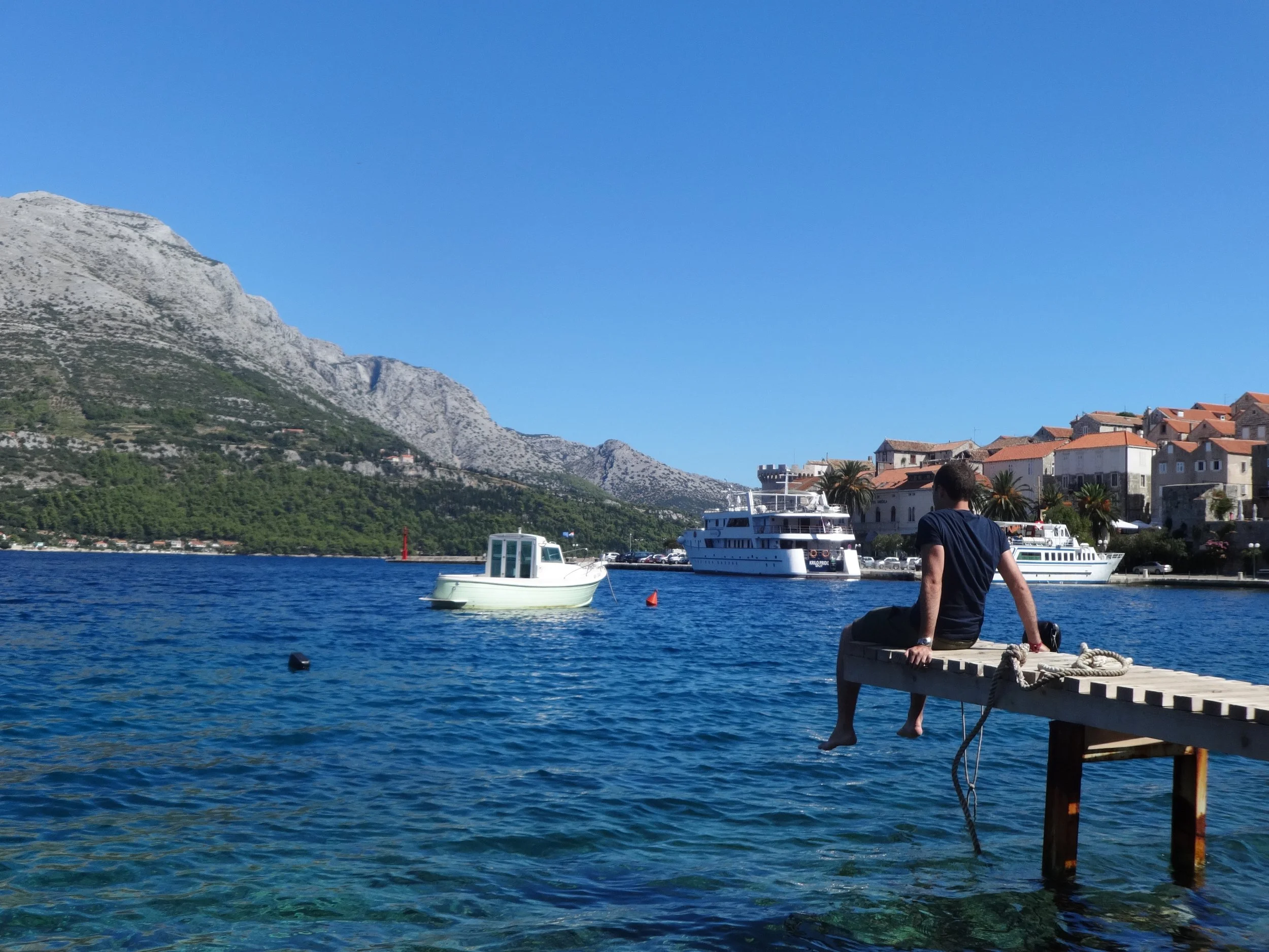 A man sitting on a dock by the water, overlooking boats and a seaside town with hills and mountains in the background, under a clear blue sky.