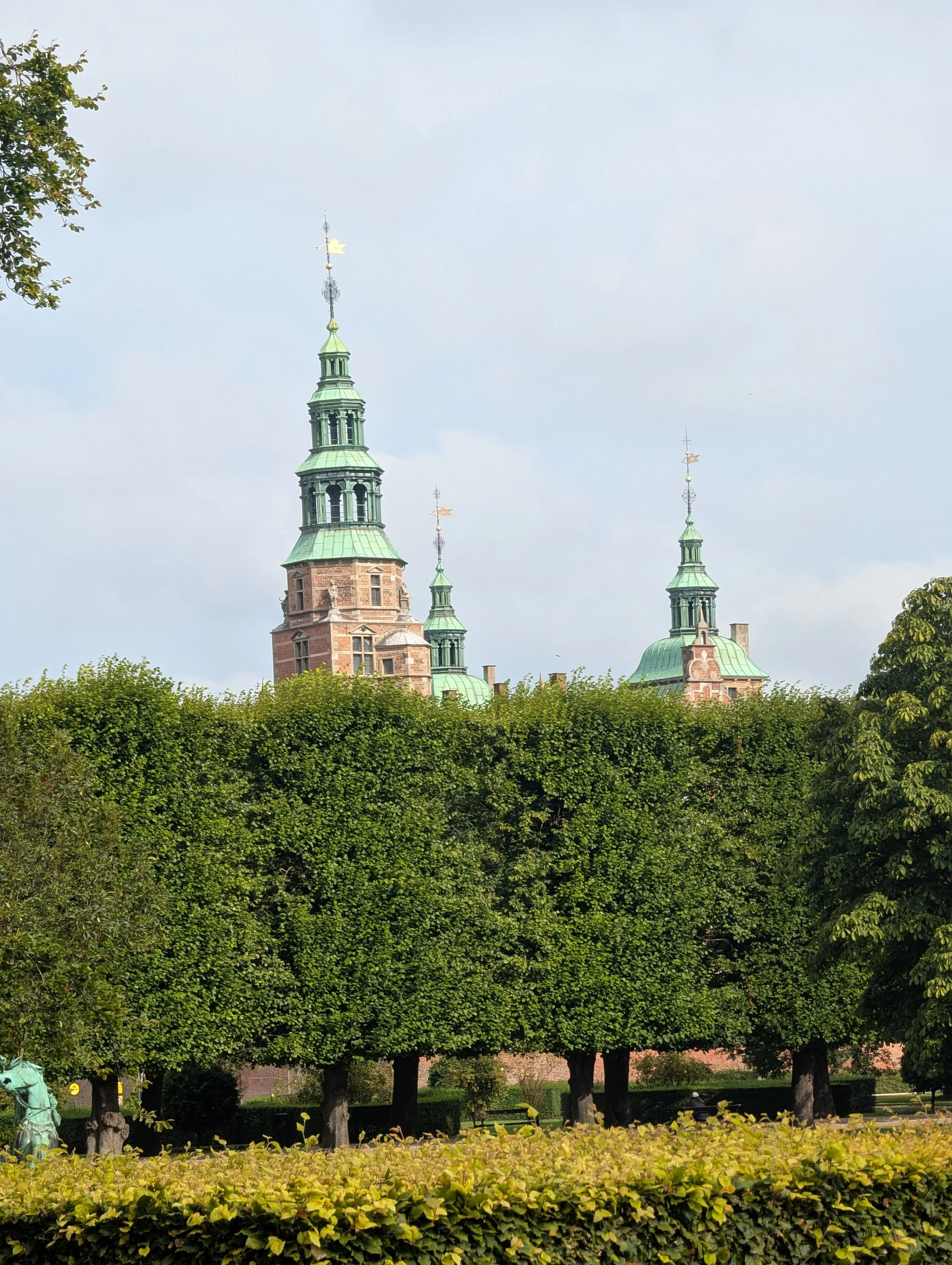 A historic building with green rooftops and spires, surrounded by trees and a hedge in the foreground.