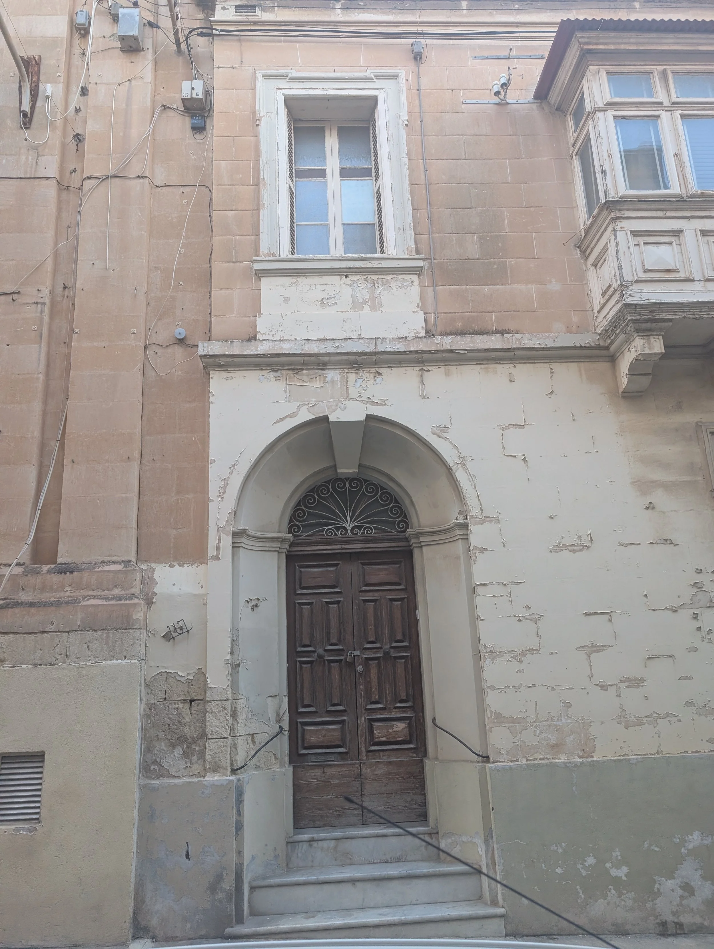 A weathered building facade featuring a wooden door, stone archway, worn white window frames, a window with shutters, and electrical wires attached to the beige stone exterior.