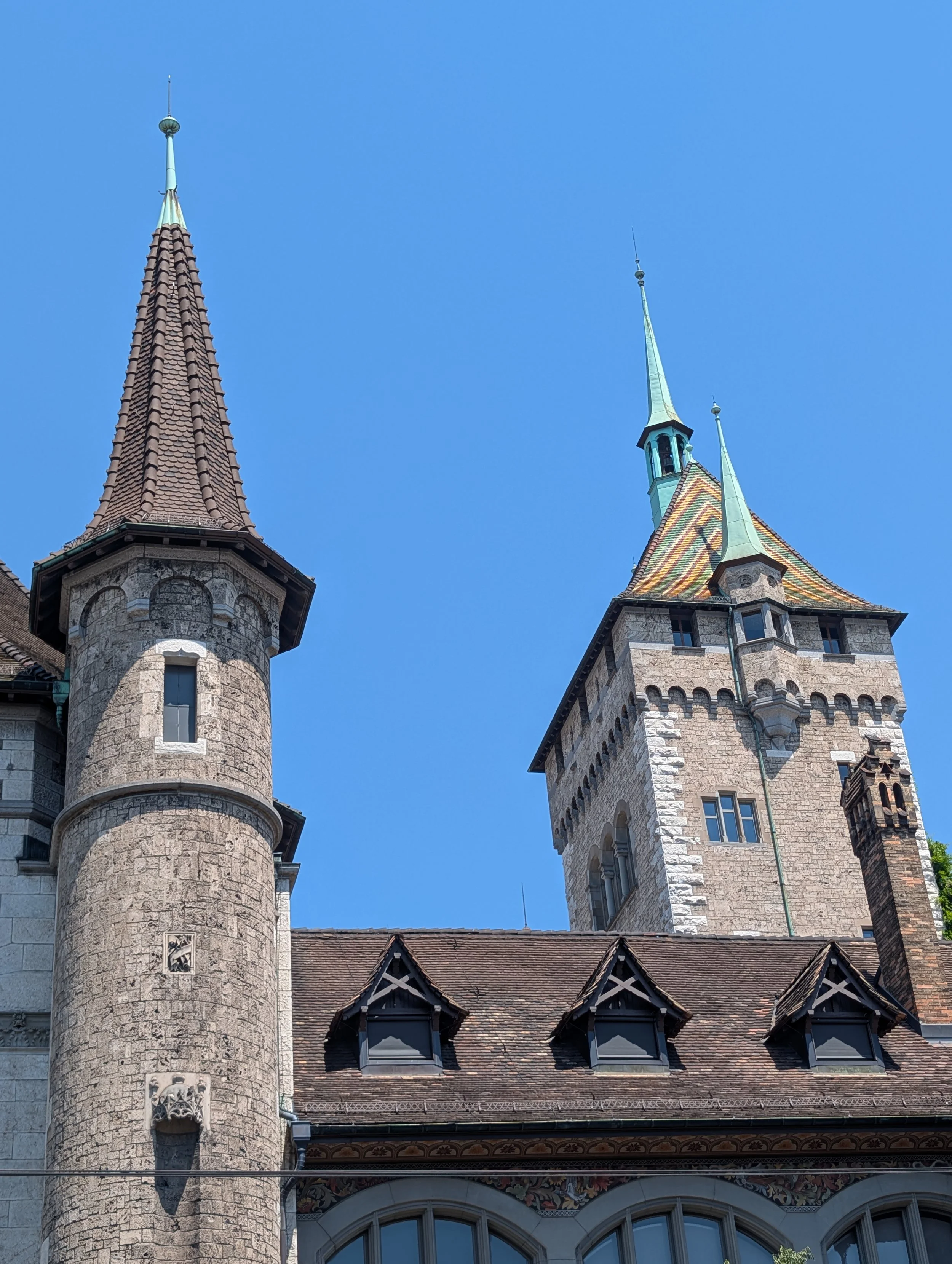 Two historic towers with pointed roofs, one with reddish tiles and the other with greenish tiles, against a clear blue sky, part of a castle or large building with intricate architecture and decorative details.