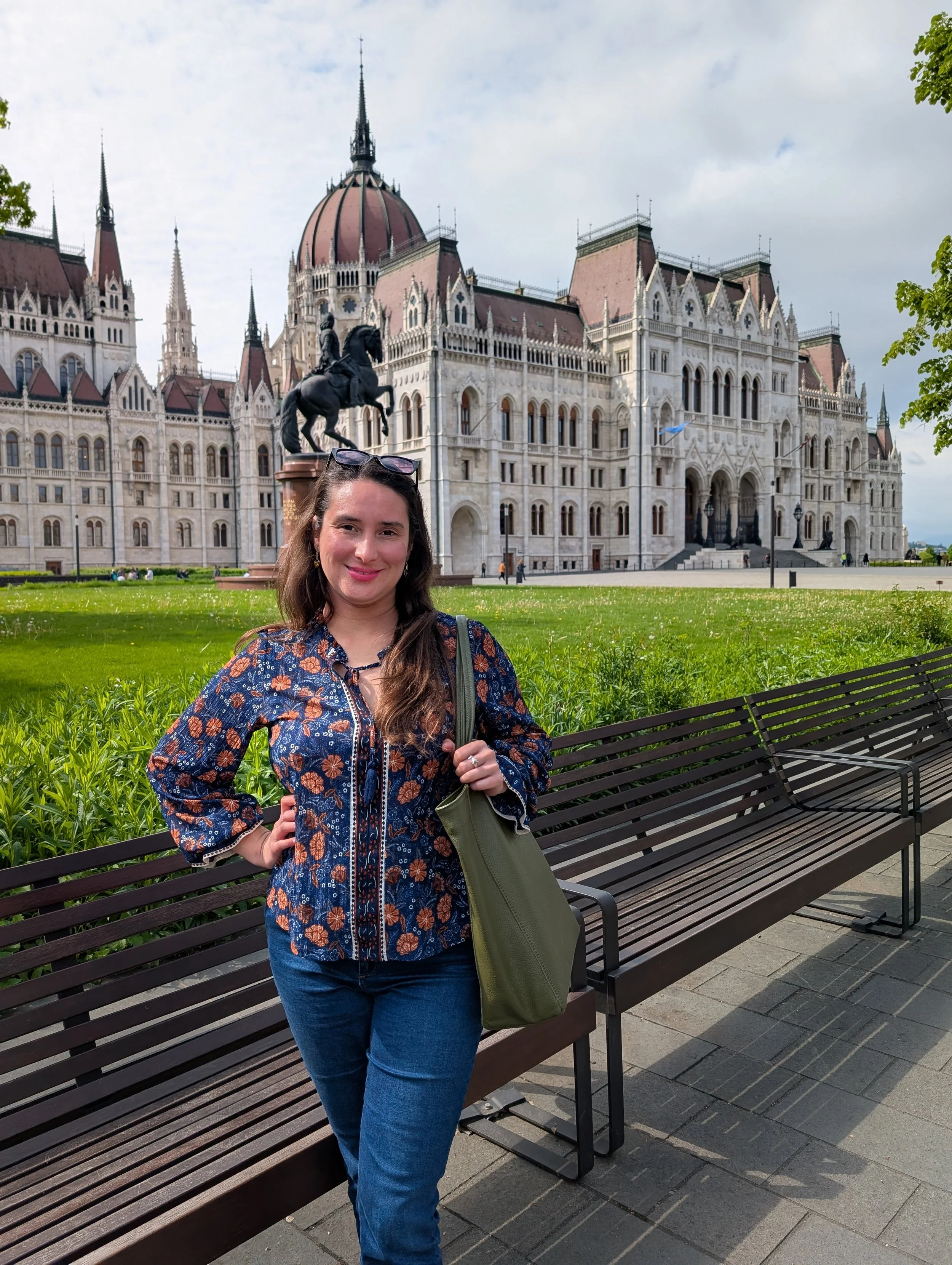 A woman with long brown hair, wearing a blue floral jacket and jeans, standing next to a park bench with a historic European-style building and a statue of a mounted knight in the background.