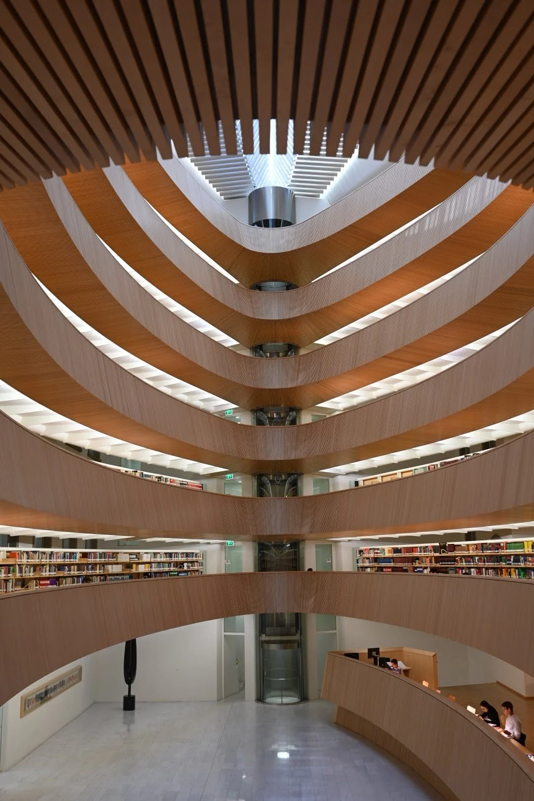 Interior of a modern multi-story library with curved wooden balconies and bookshelves, viewed from the ground floor looking upward.