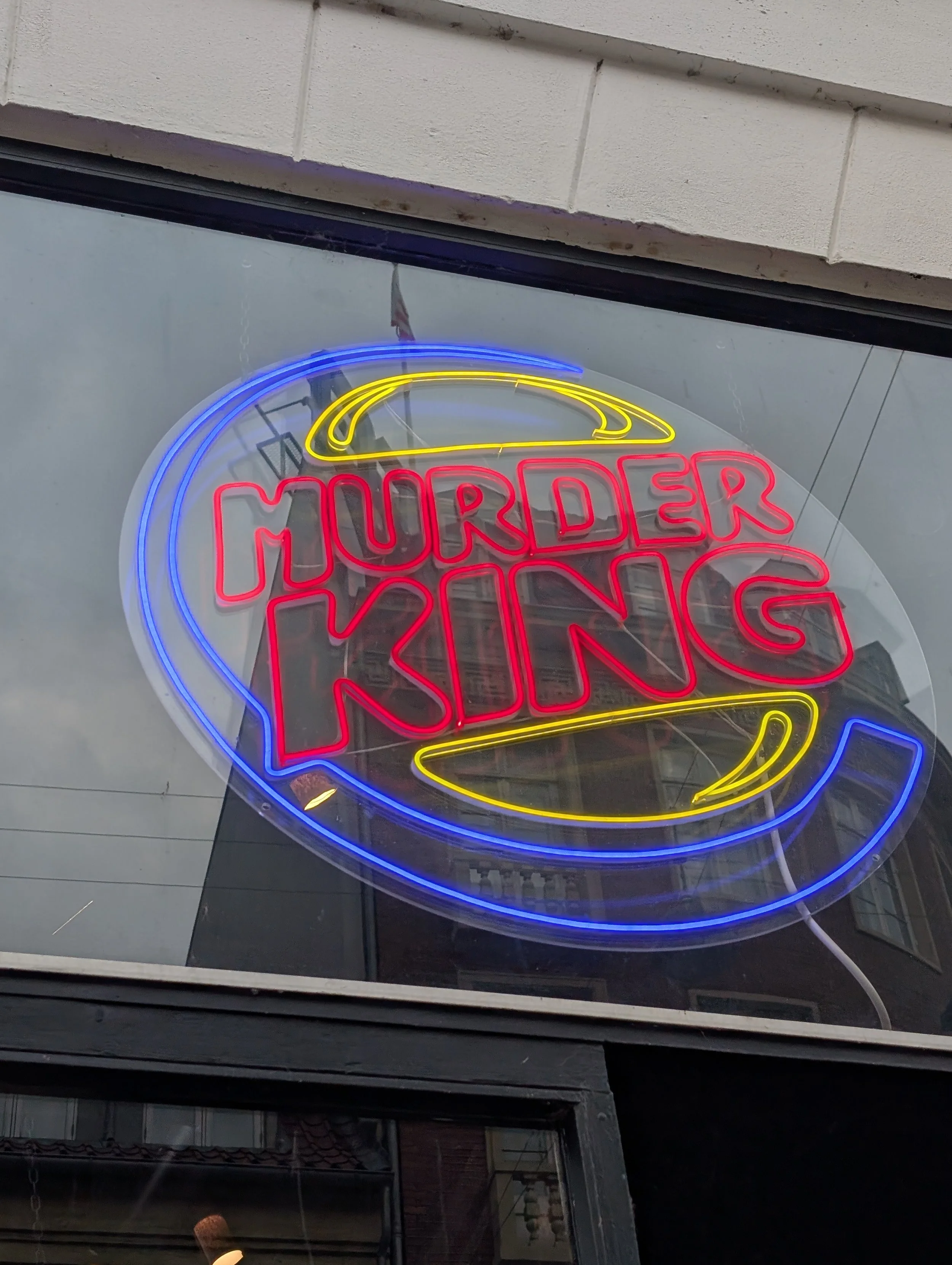 Neon sign with a yellow fried egg and red letters that read 'Murder King' in a window display at night.