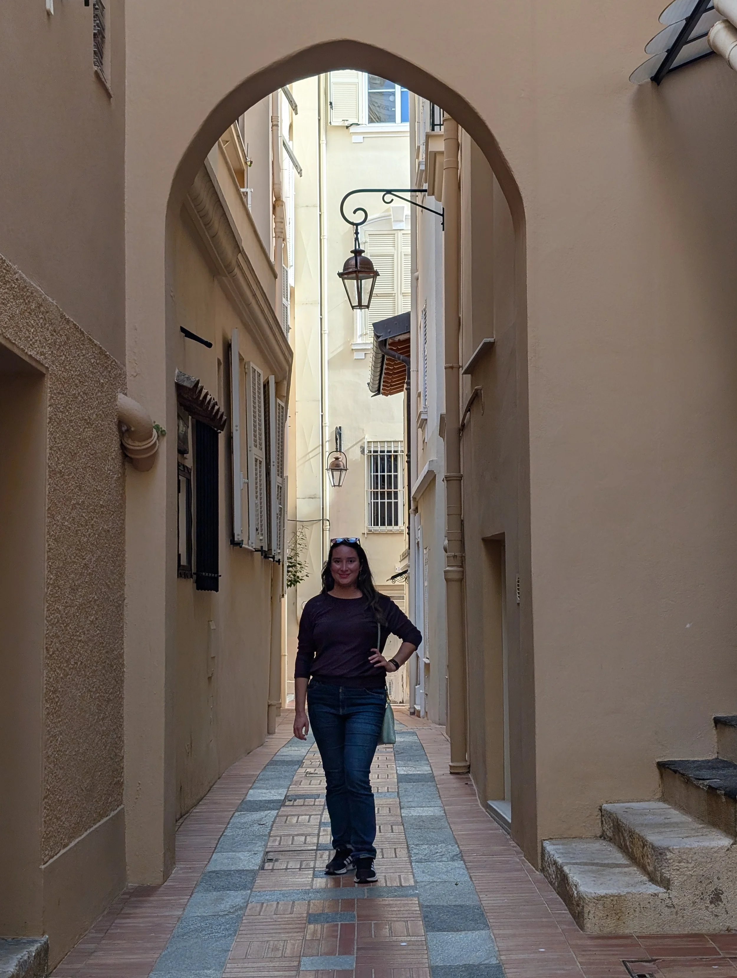 A woman standing in a narrow alleyway with beige and tan walls, arched entry, decorative black lantern hanging, and a tile path.
