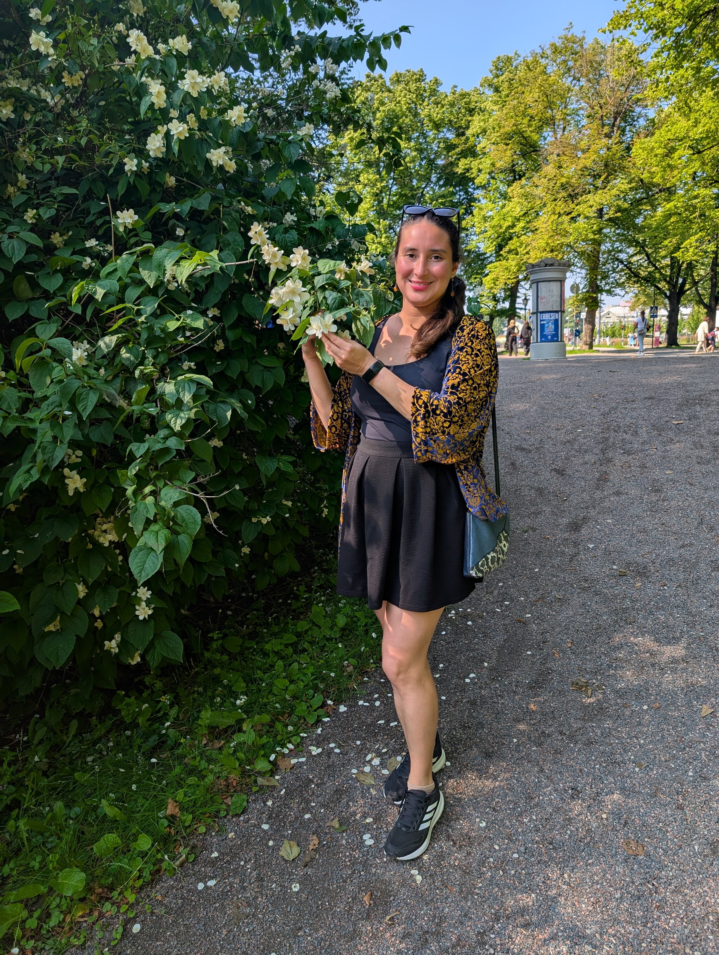 A woman standing outdoors on a gravel path, smiling, holding white flowers from a bush, with trees and a park in the background on a sunny day.