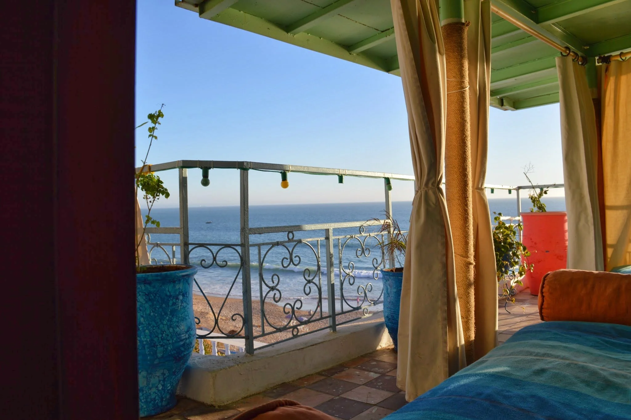 View from a balcony overlooking the ocean, with a string of colourful lights hanging, curtains partially drawn, and potted plants on the balcony.