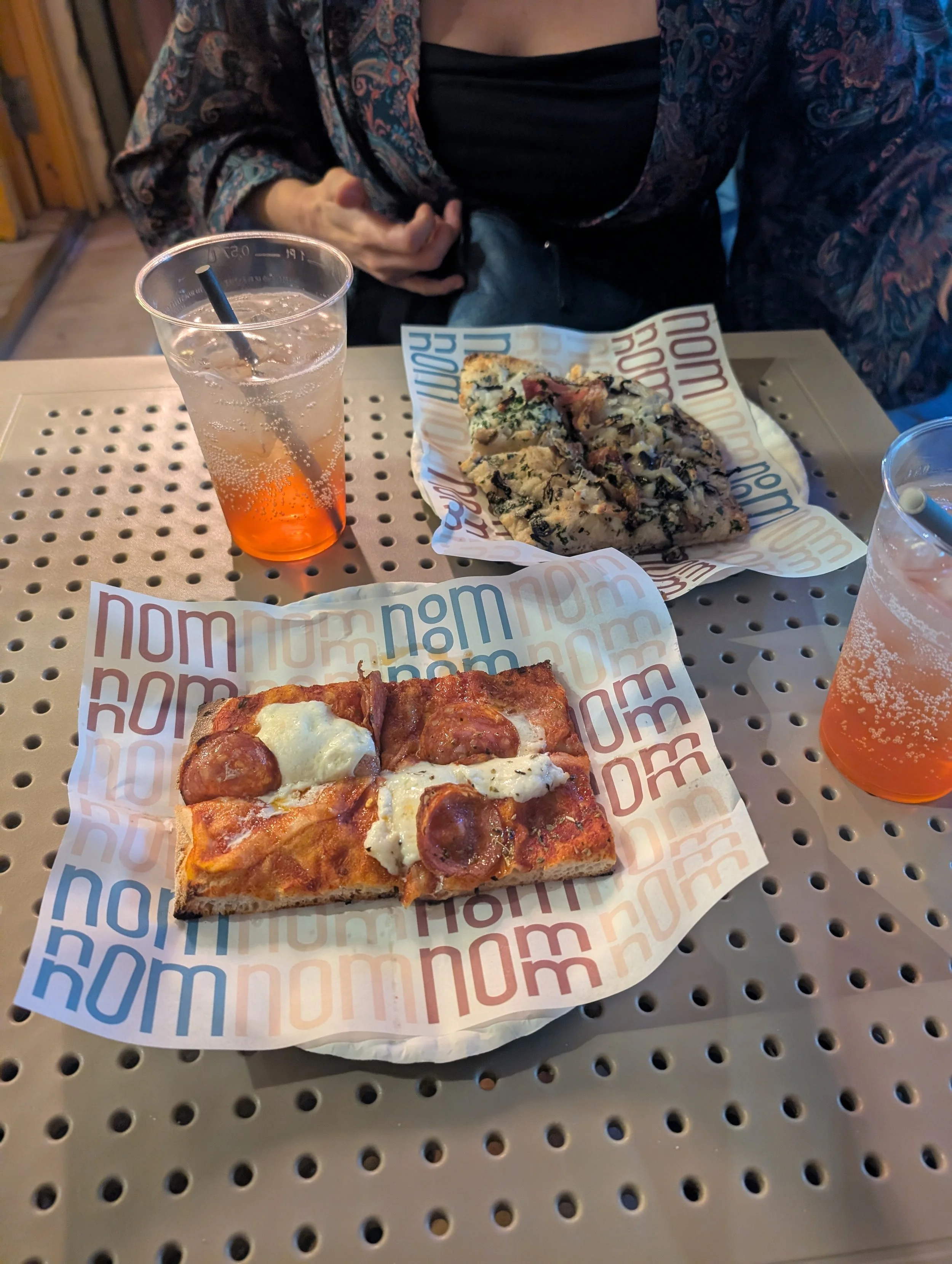 A table with two slices of pizza and two glasses of soda. One pizza slice has pepperoni and cheese, the other has various toppings including what appears to be mushrooms and herbs. The drinks are iced, with one having a black straw. The table surface