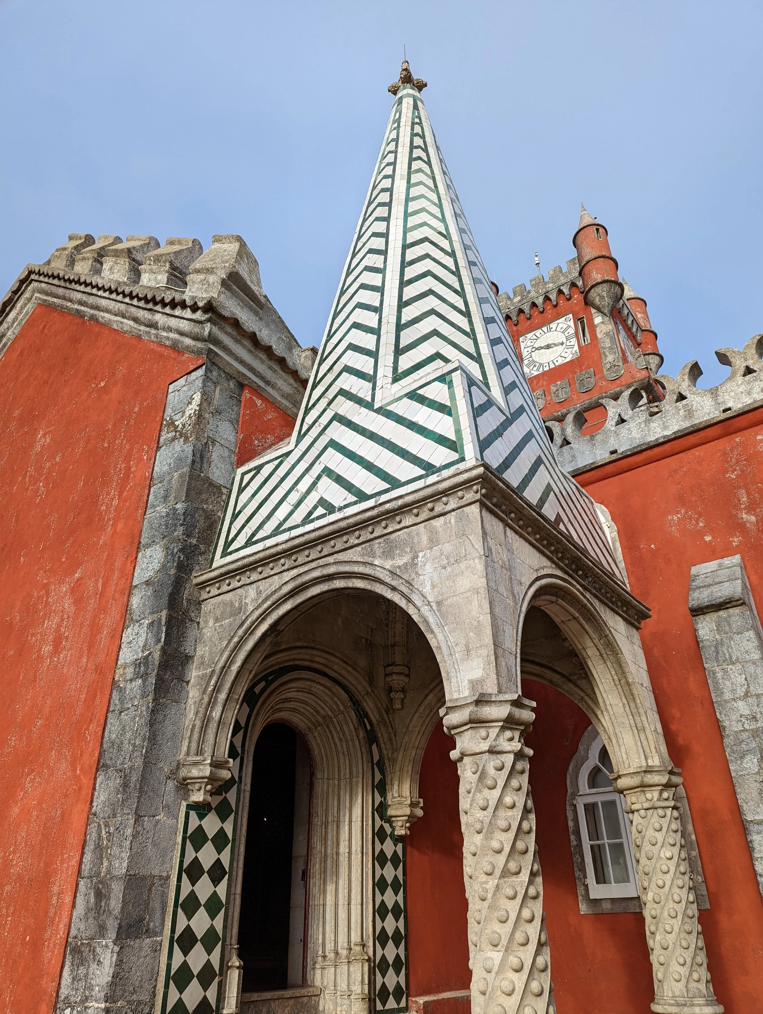 Low-angle view of a church entrance with gothic arches, among orange walls, a chevron-patterned spire, and a clock tower against a clear blue sky.